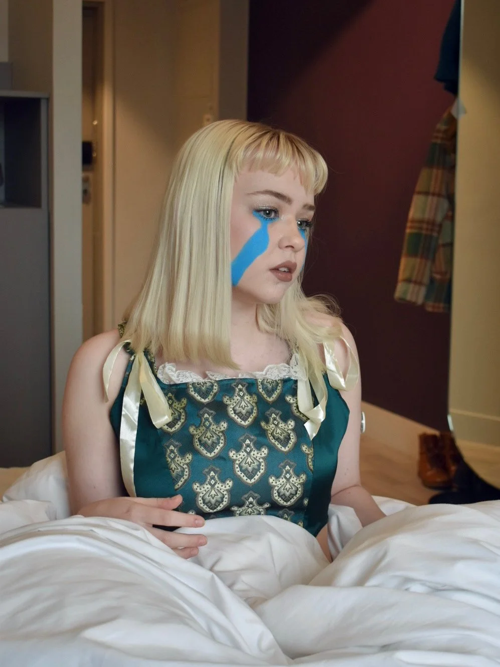 Greta, a young white blonde woman wearing a dark green corset top with gold details and white satin bows, is sitting on a white bed and looking away. Two bright blue big tears are painted across her cheeks.