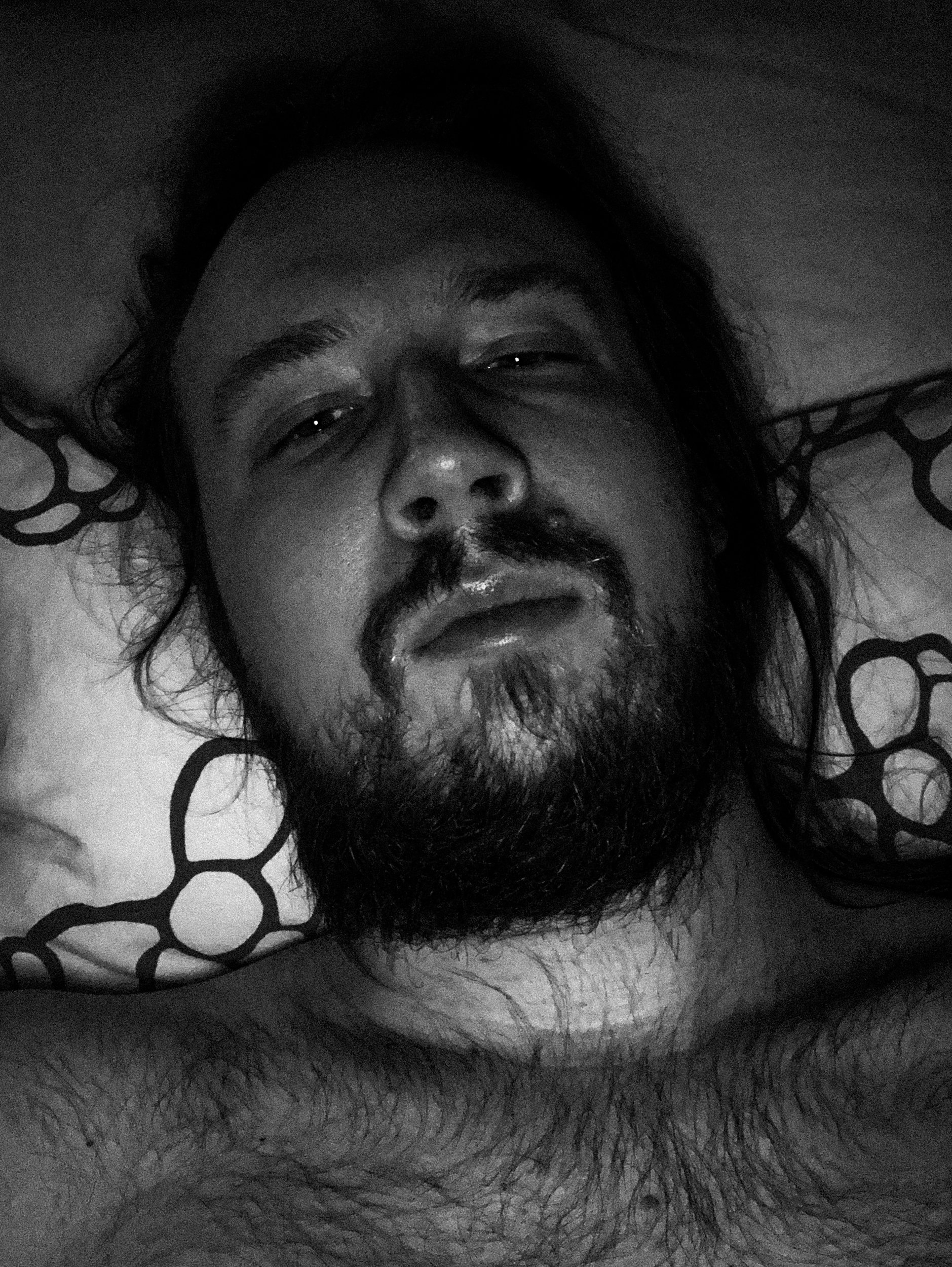 A black-and-white selfie photo of Niko in a dark bedroom; he’s looking directly at the camera while lying in bed. He has a messy beard and long hair.