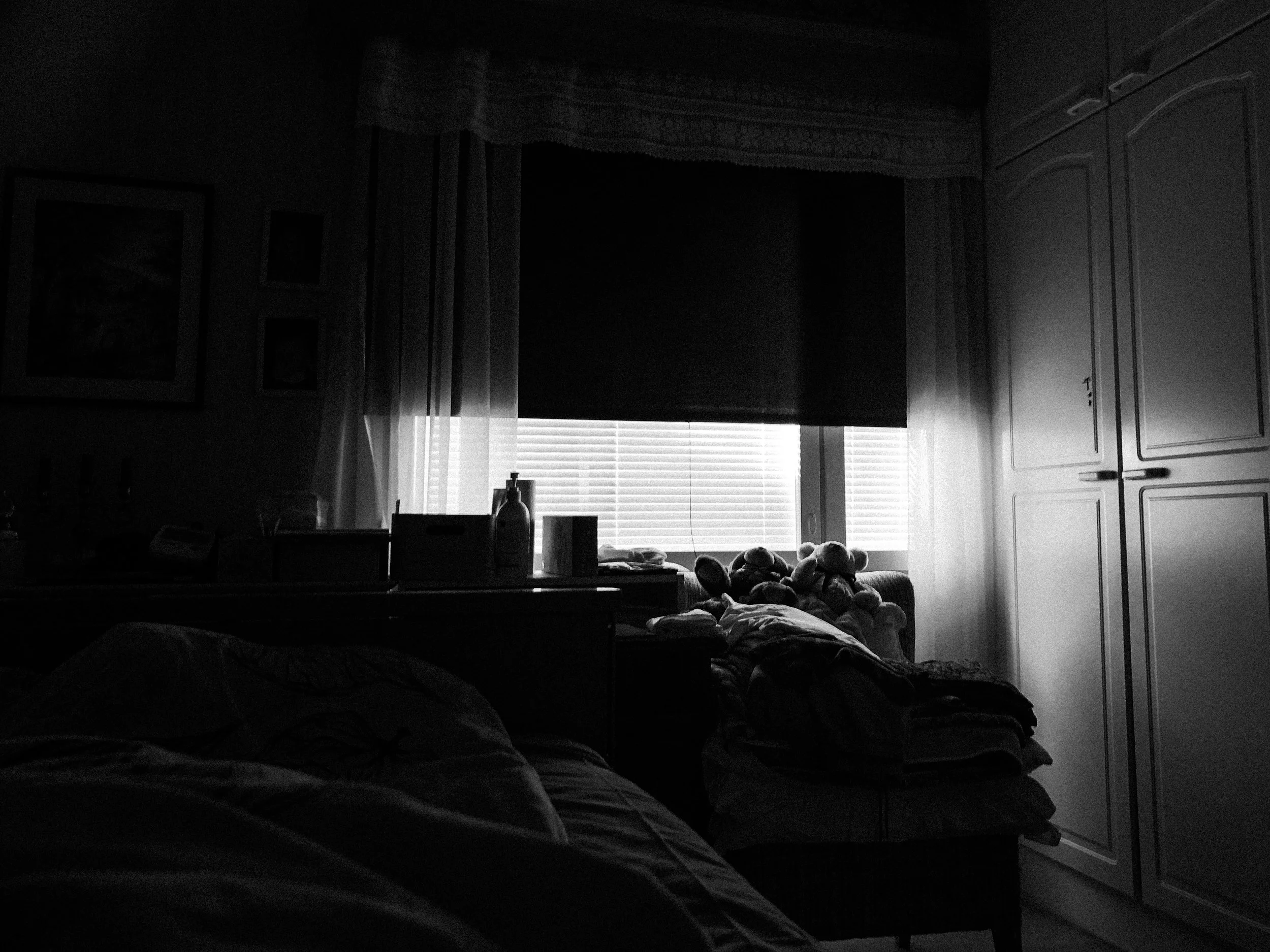 A black-and-white high contrast photo of a dark bedroom window; the blinds are fully closed, the blackout curtain is lifted a bit to let more light in.