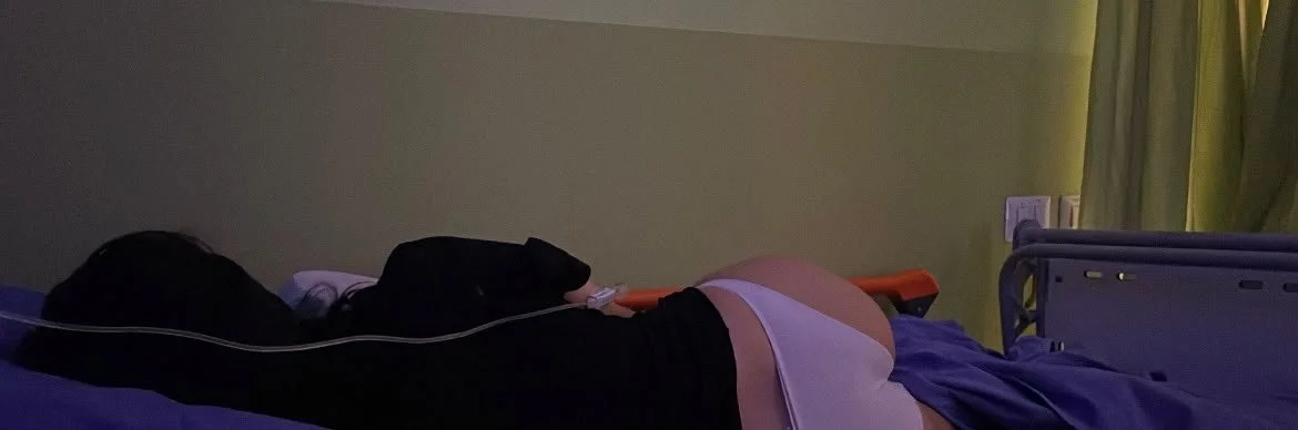 A photograph of a young woman lying in a hospital bed in a dimly lit room, facing away from the camera. She has long dark hair and wears a black t-shirt and white underwear, and has an IV tube connected to her arm.