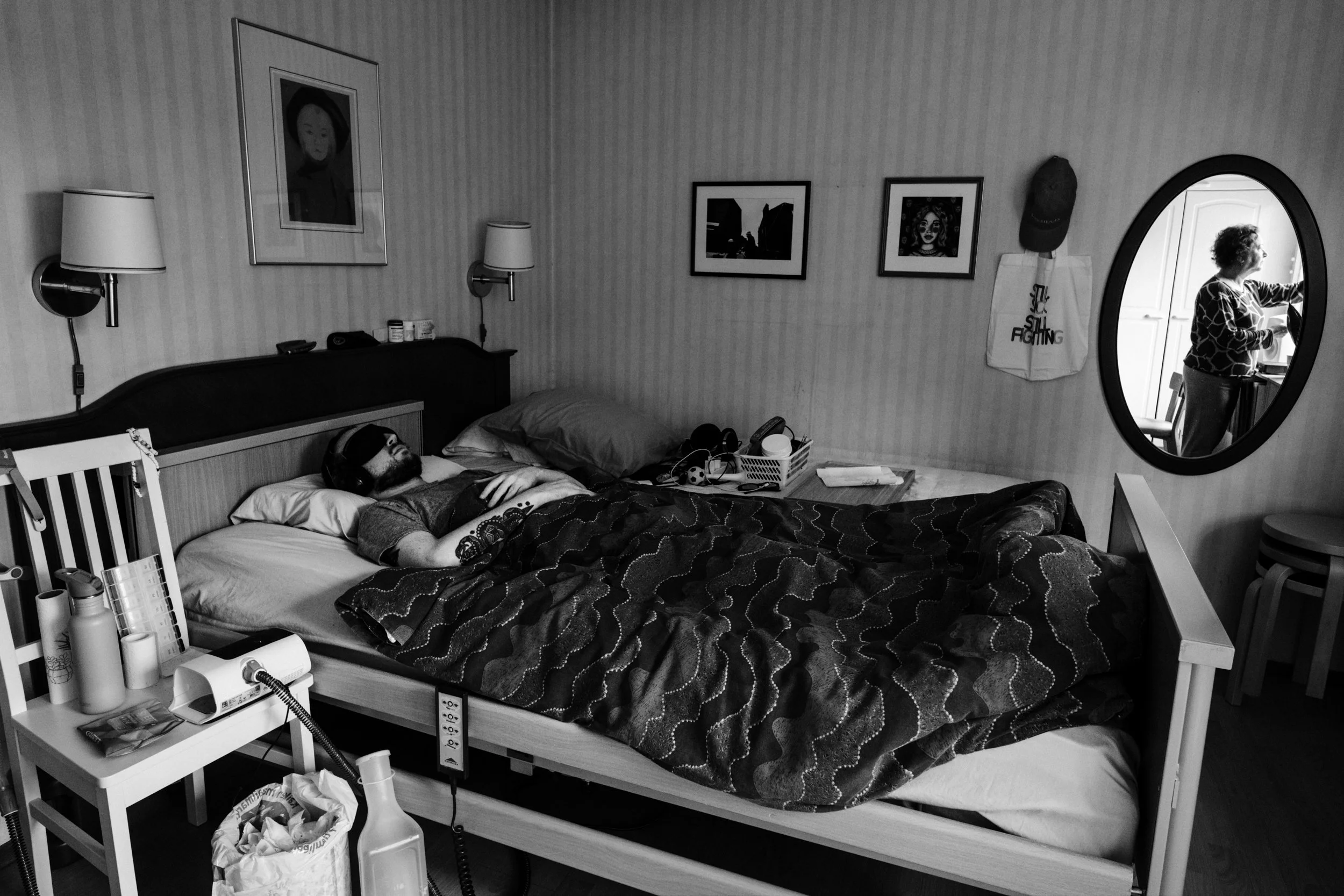 A black-and-white photo of a man lying on a hospital bed, hands crossed on his chest, under a duvet, and wearing an eyemask and earmuffs. The room has framed pictures and personal belongings. A mirror reflects a woman closing the curtains.