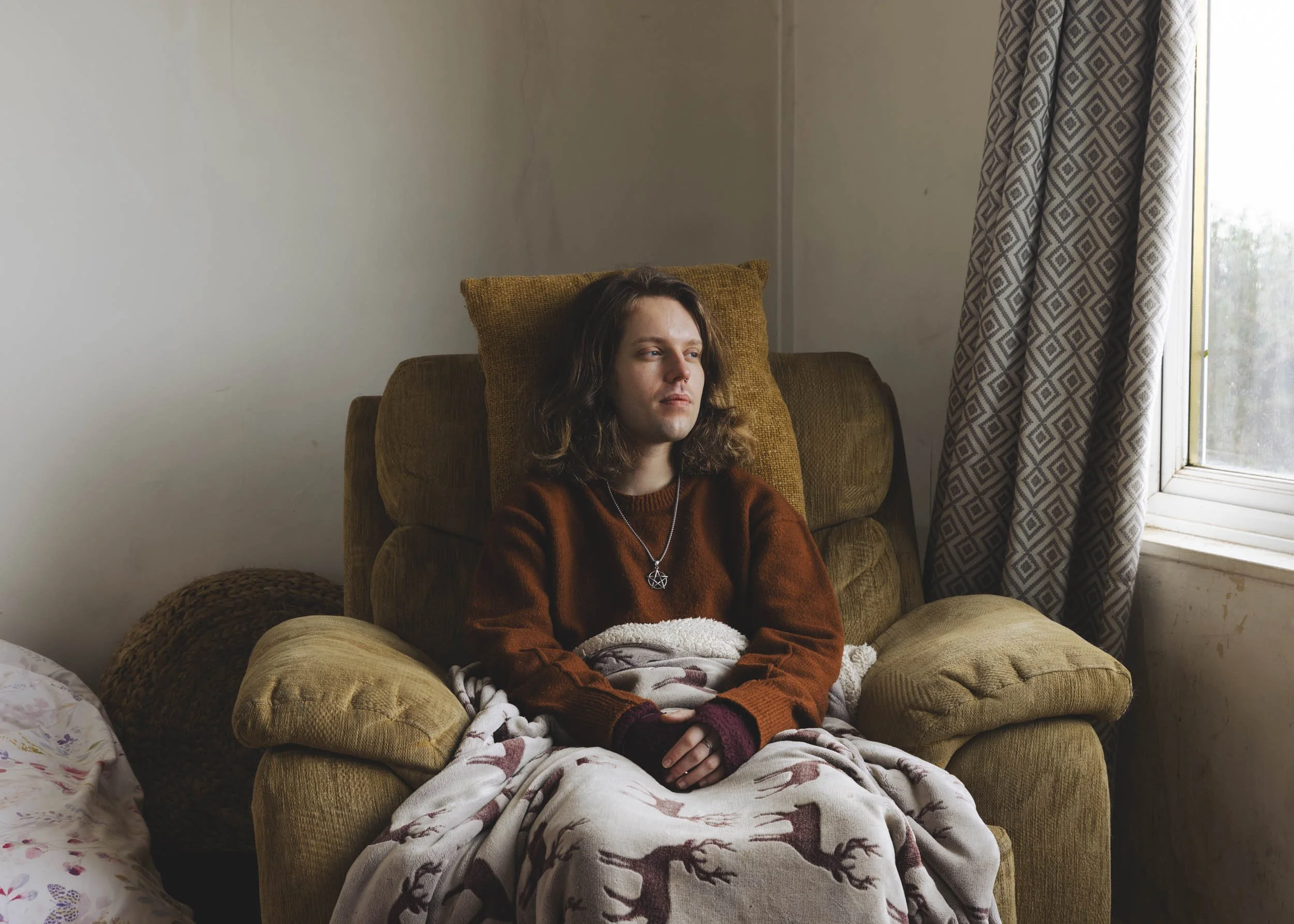 A photograph of Jack, a fair-skinned man with wavy, brown, shoulder-length hair, looking out the window to his left. He sits in a large lounge chair and has a blanket on his lap. On the walls, there is water damage and specks of black.