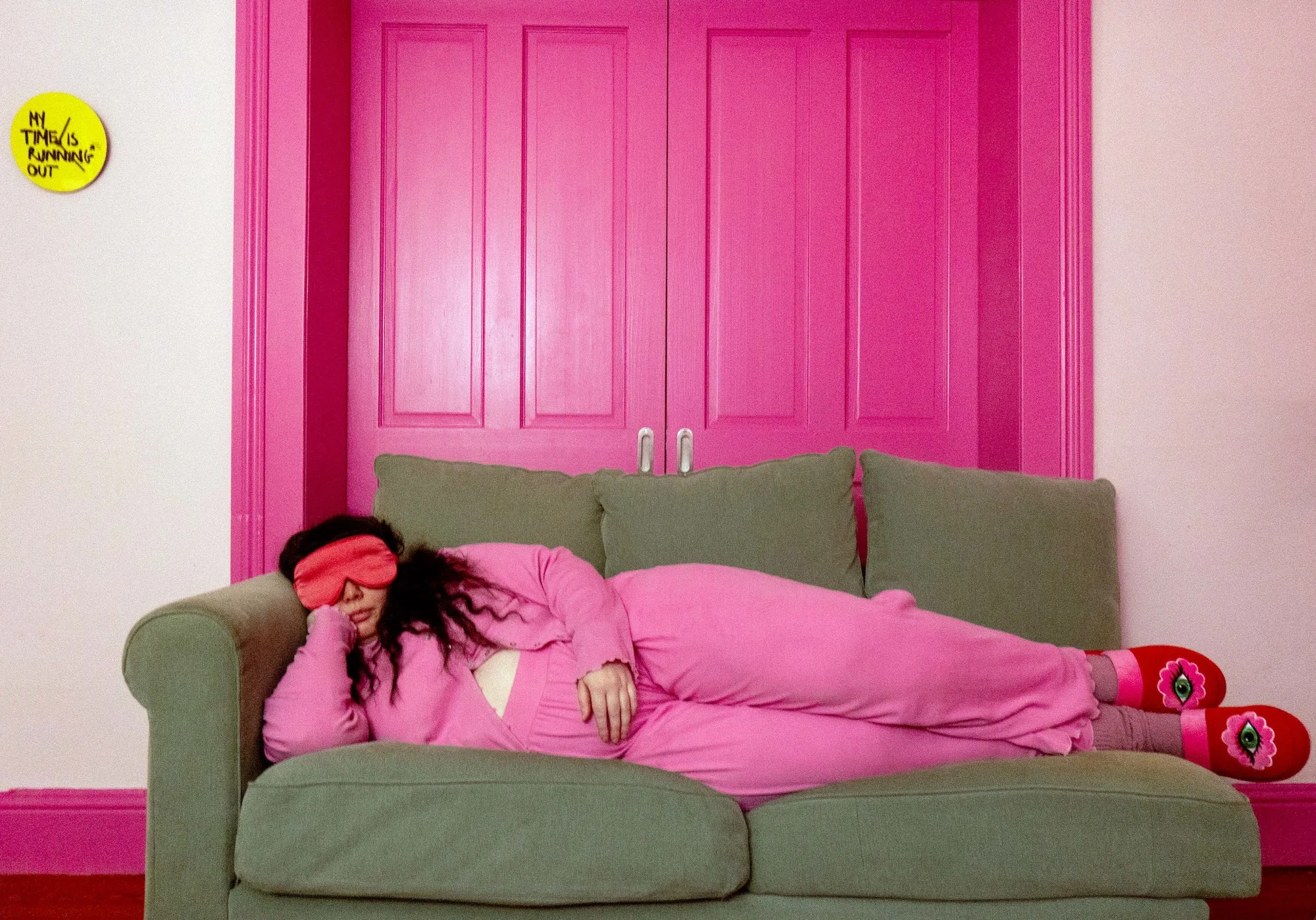 A photograph of a woman in pink lounge wear and an eyemask lying on a green sofa. In the background is a set of pink doors, and on the wall a yellow clock that says ‘my time is running out’.