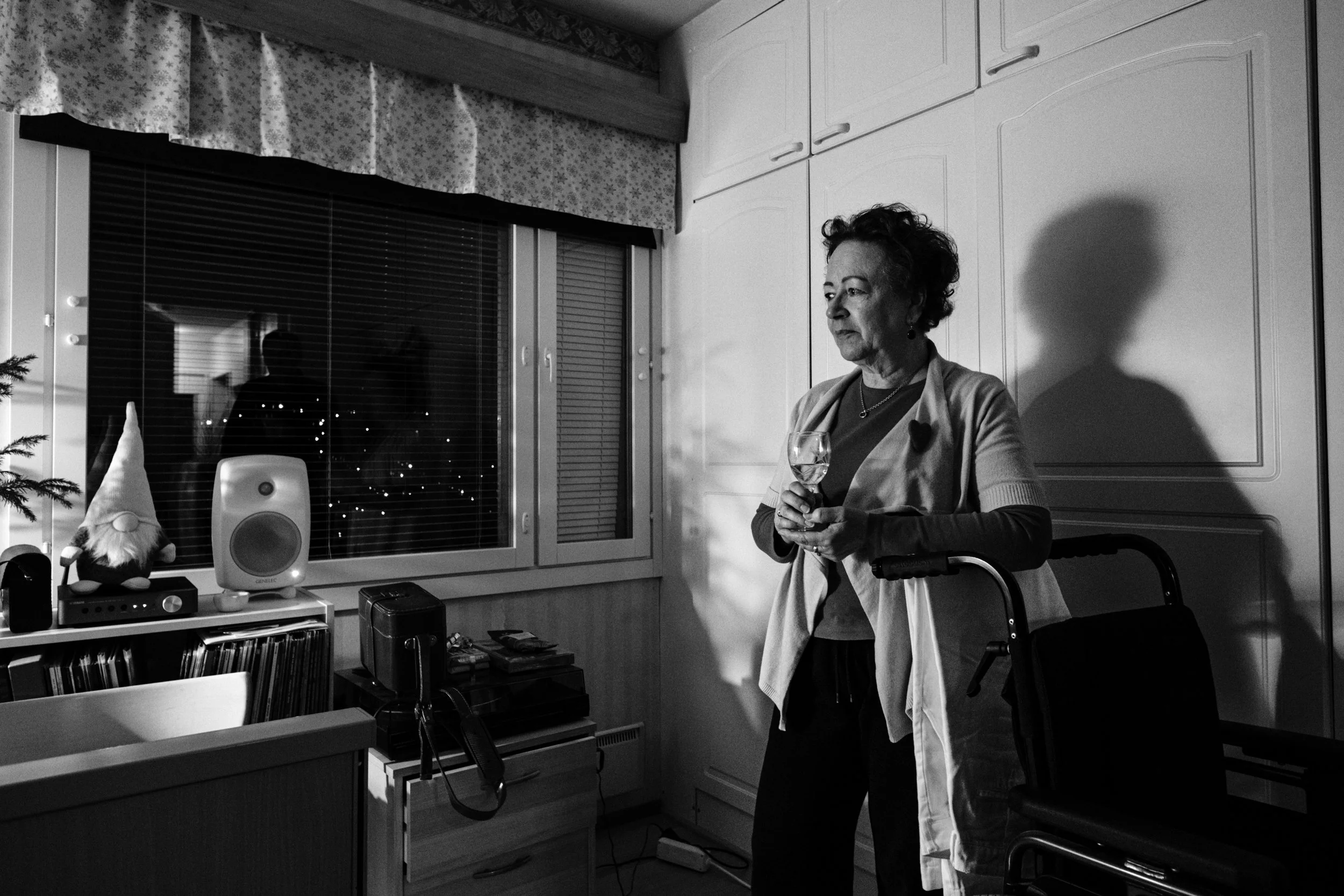 A black-and-white photo of Päivi, a woman with short, curly hair, standing in a dimly lit room holding a wine glass. She leans on a wheelchair. Her shadow falls on white cabinets. A window with blinds reflects another silhouette.