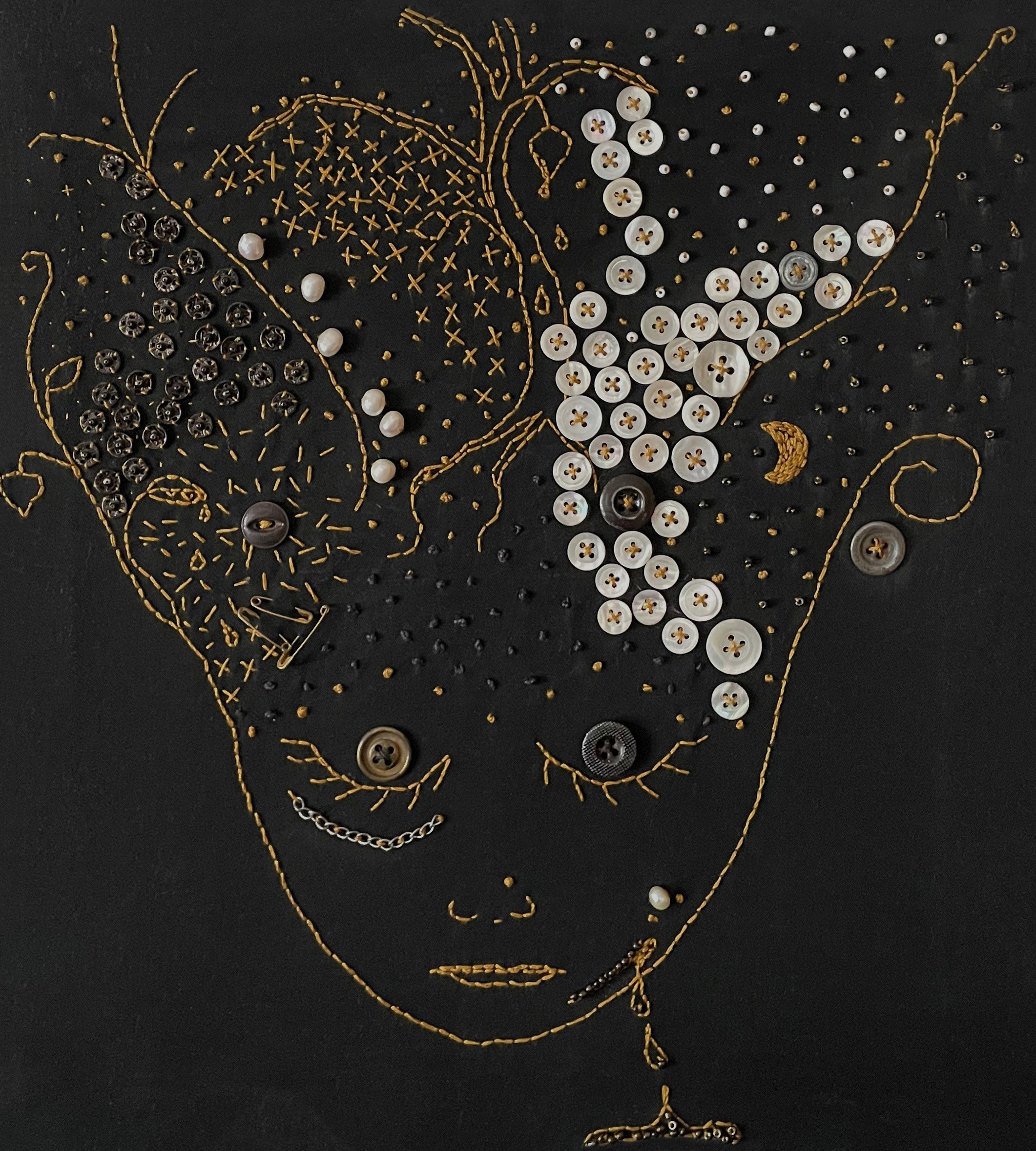 The head of a woman embroidered in yellow stitching on black fabric. She has full hair, both dark and light, where a small tree, floral stems, and a small moon grow. She has buttons on her eyelids, and on her face is a bleeding wound.