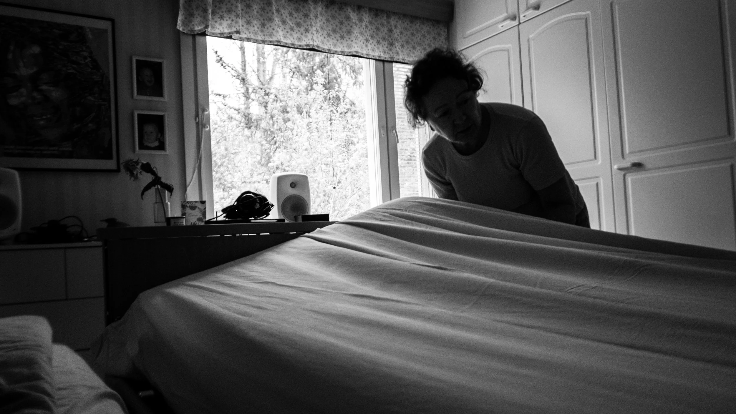 A black-and-white photo of Päivi making a bed, bending slightly to tuck a sheet under the mattress. A window in the background lets in natural light, illuminating a dresser with personal items.