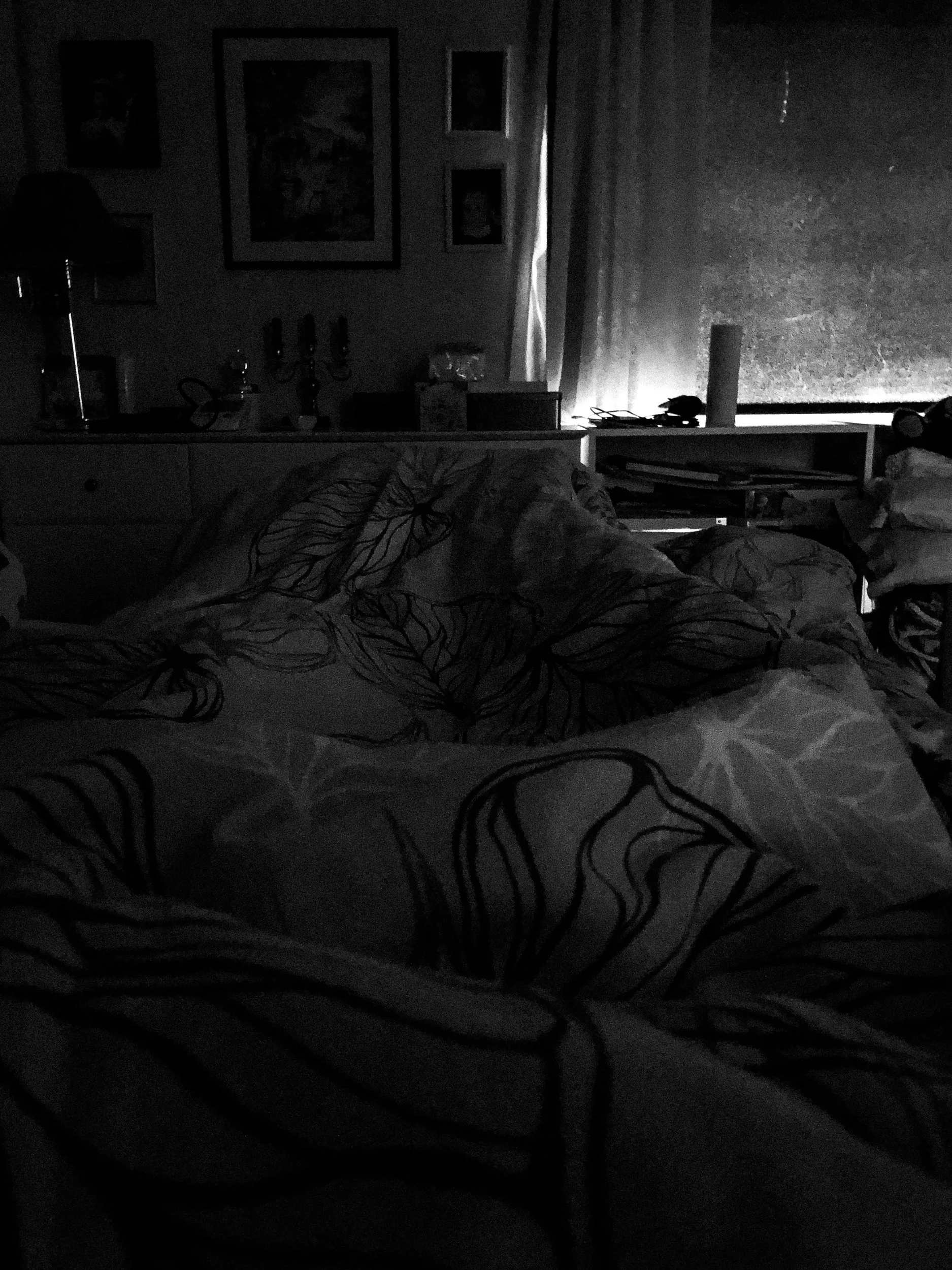 A black-and-white photo of Niko’s point of view showing his pitch-black bedroom while he’s lying in bed. Only a small ray of light peeks under the curtains.