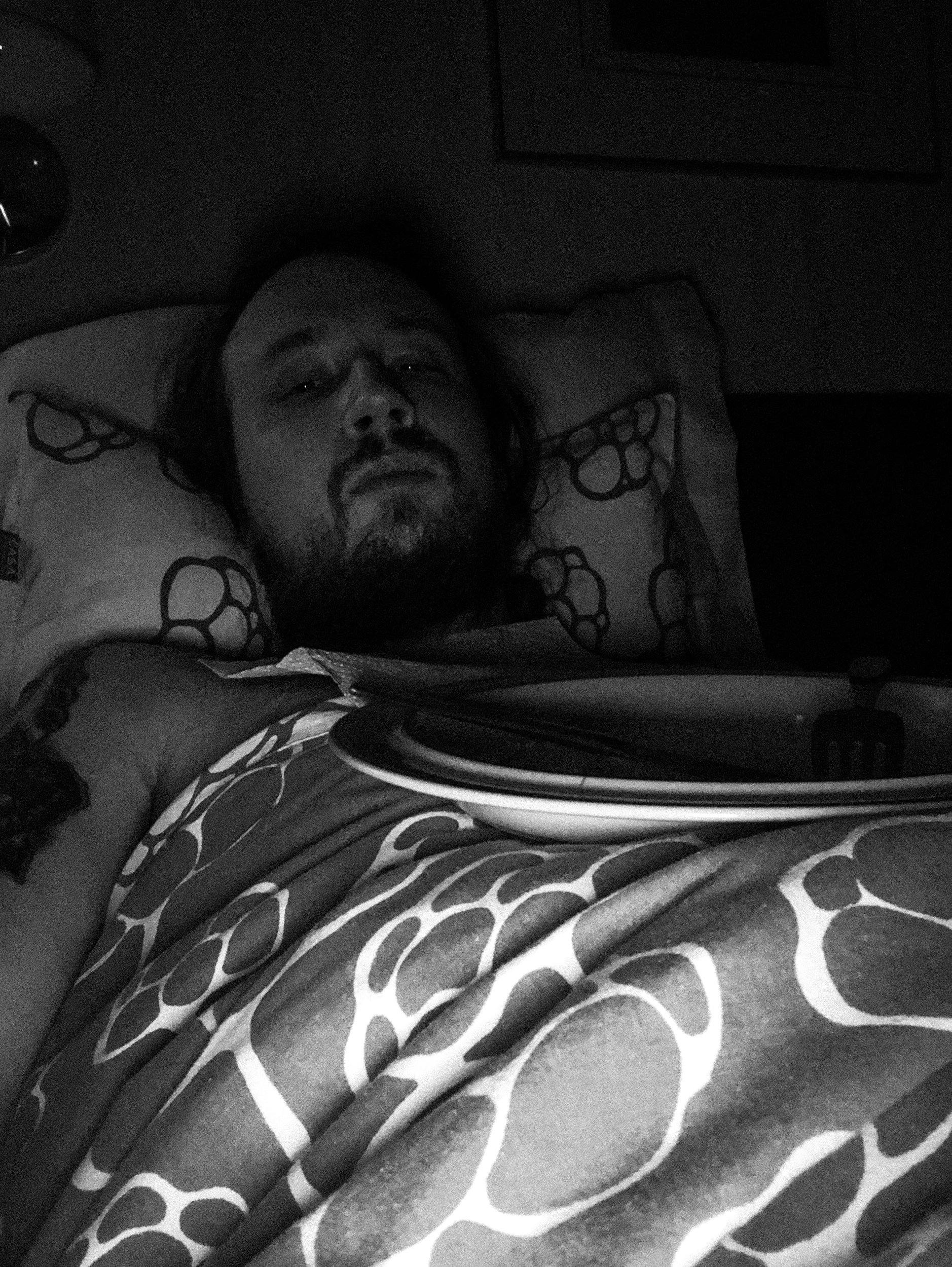 A black-and-white selfie photo of Niko in a dark bedroom; he’s looking directly at the camera while lying in bed. He has a messy beard and long hair. A finished plate of food is lying on his stomach.