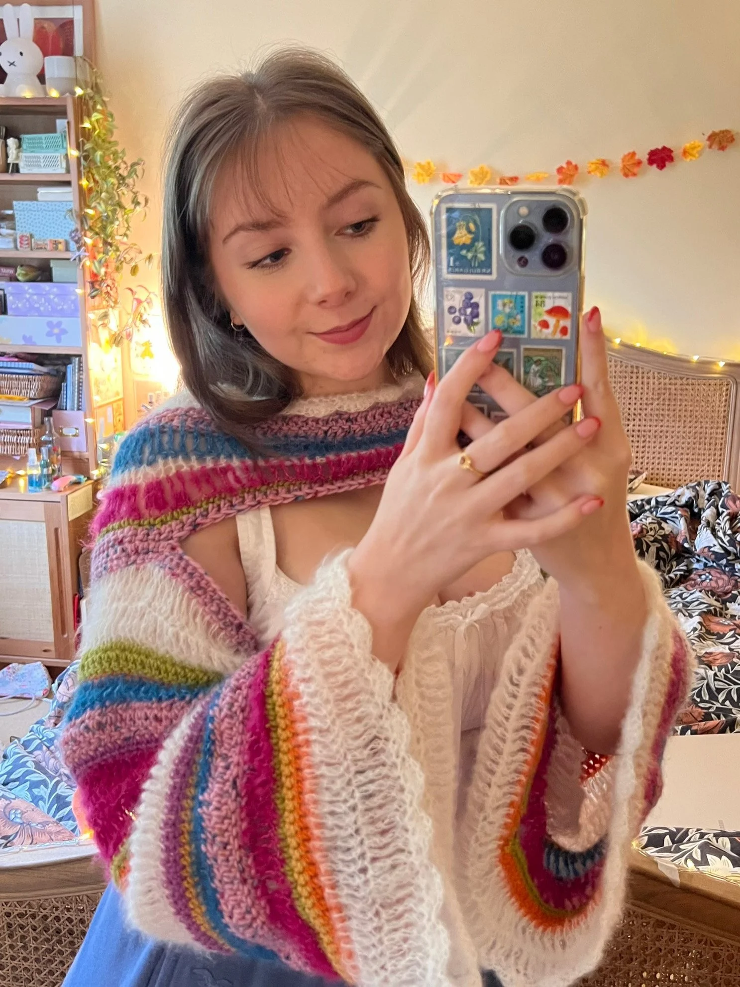 Emily, a young white woman with wavy brown hair, wearing a colorful striped crochet top, is in a bedroom, holding a phone, and smiling lightly.
