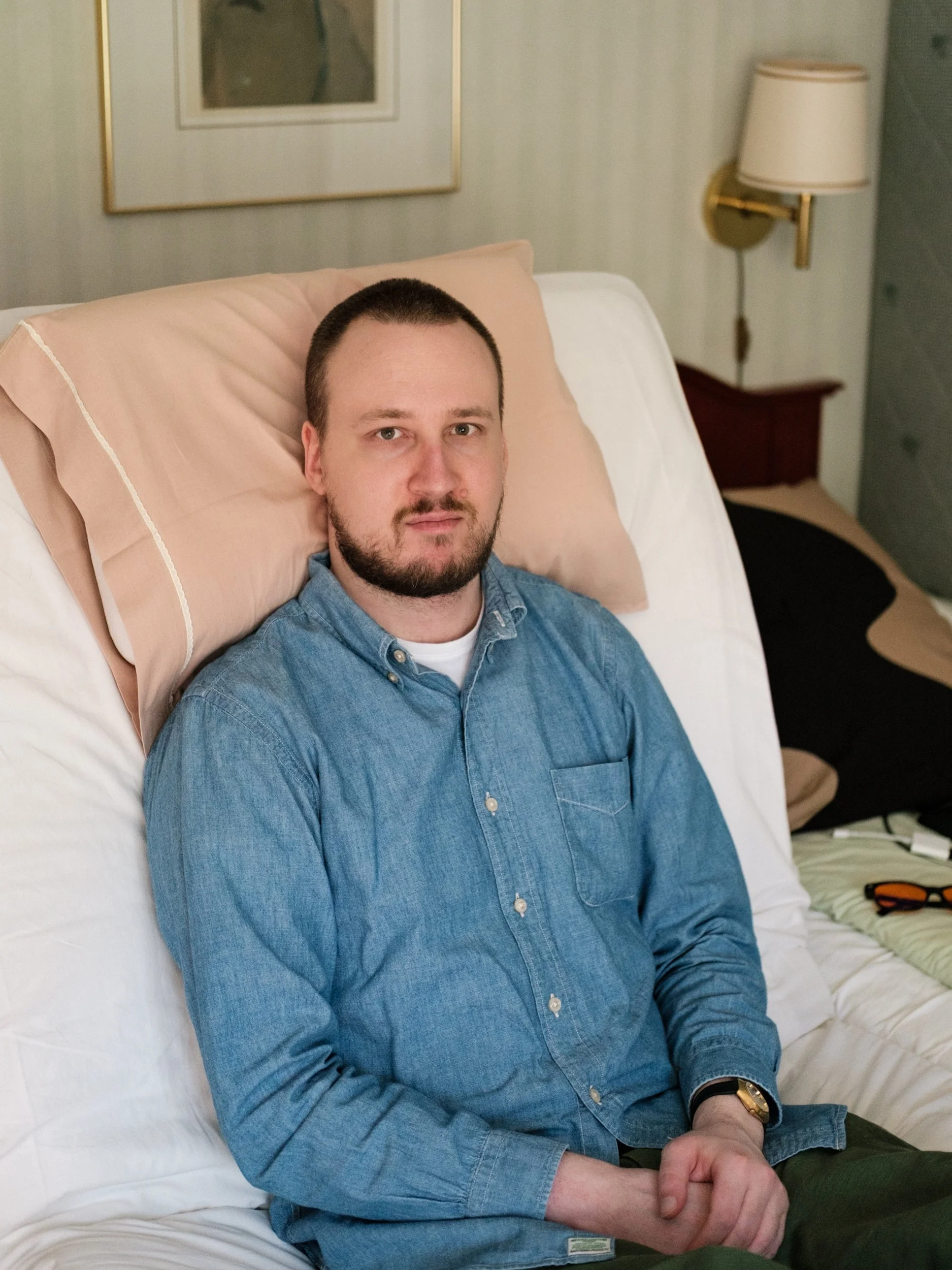 Niko, a white man with short dark hair and a beard sits upright in a hospital bed with a neutral expression. He is wearing a denim button-up shirt and a gold watch on his wrist. The setting is a bedroom.