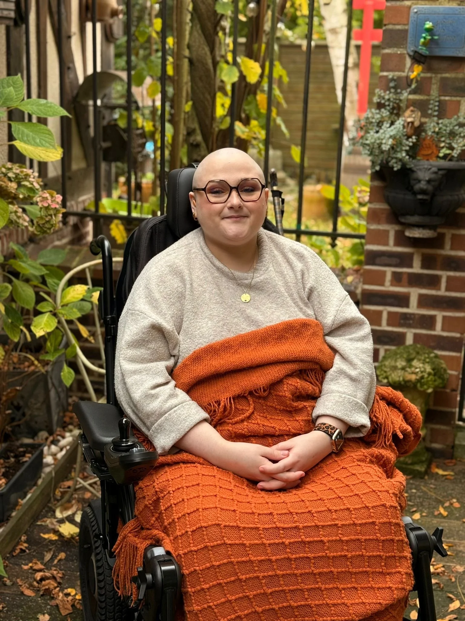 Alicia, a smiling young white woman with glasses and a shaved head, is seated in a power wheelchair. She has an orange blanket over her lap and is in a garden setting with brick walls and foliage in the background.