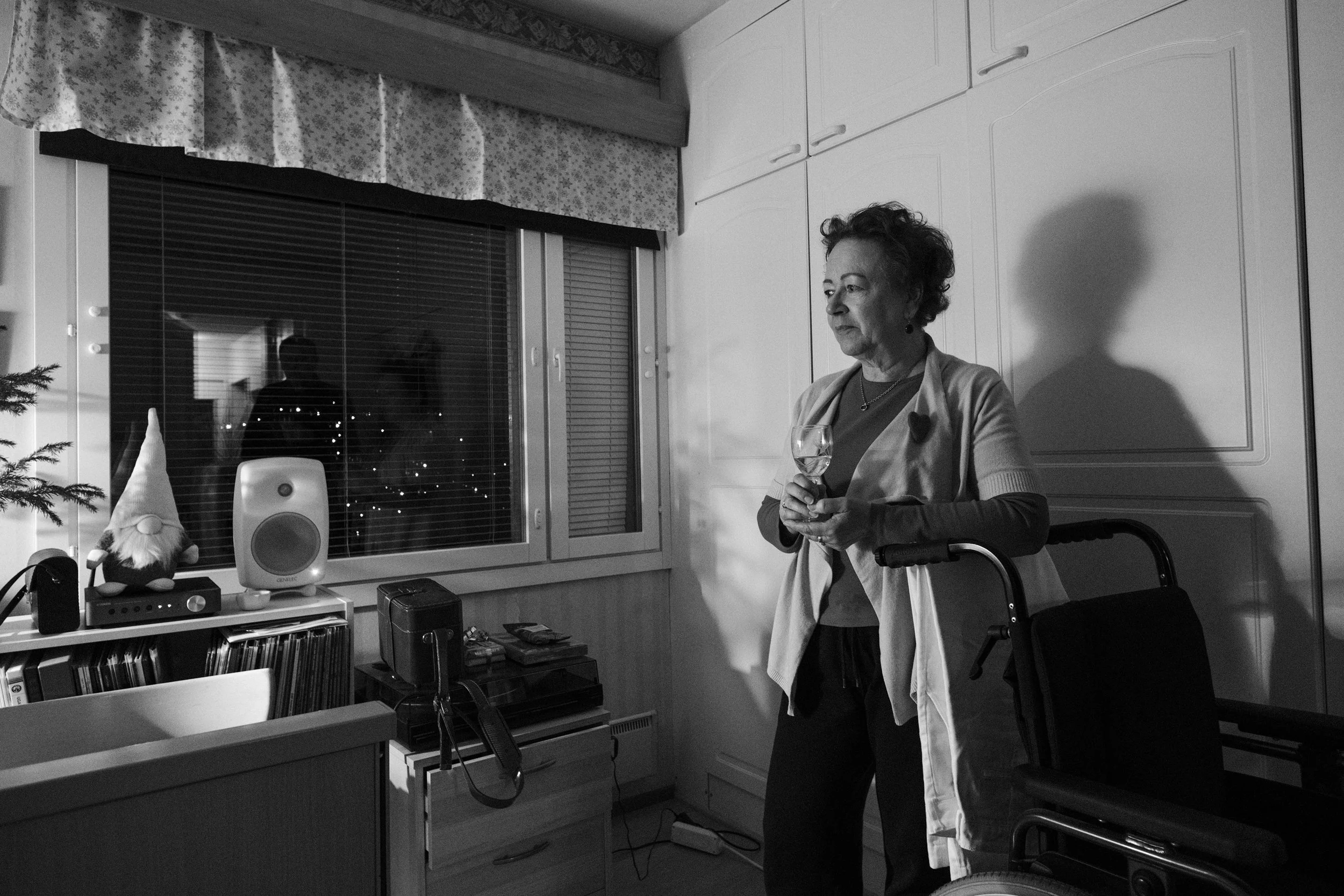A black and white photo of Päivi, a woman with short, curly hair, standing in a dimly lit room holding a wine glass. She leans on a wheelchair. Her shadow falls on white cabinets. A window with blinds reflects another silhouette.