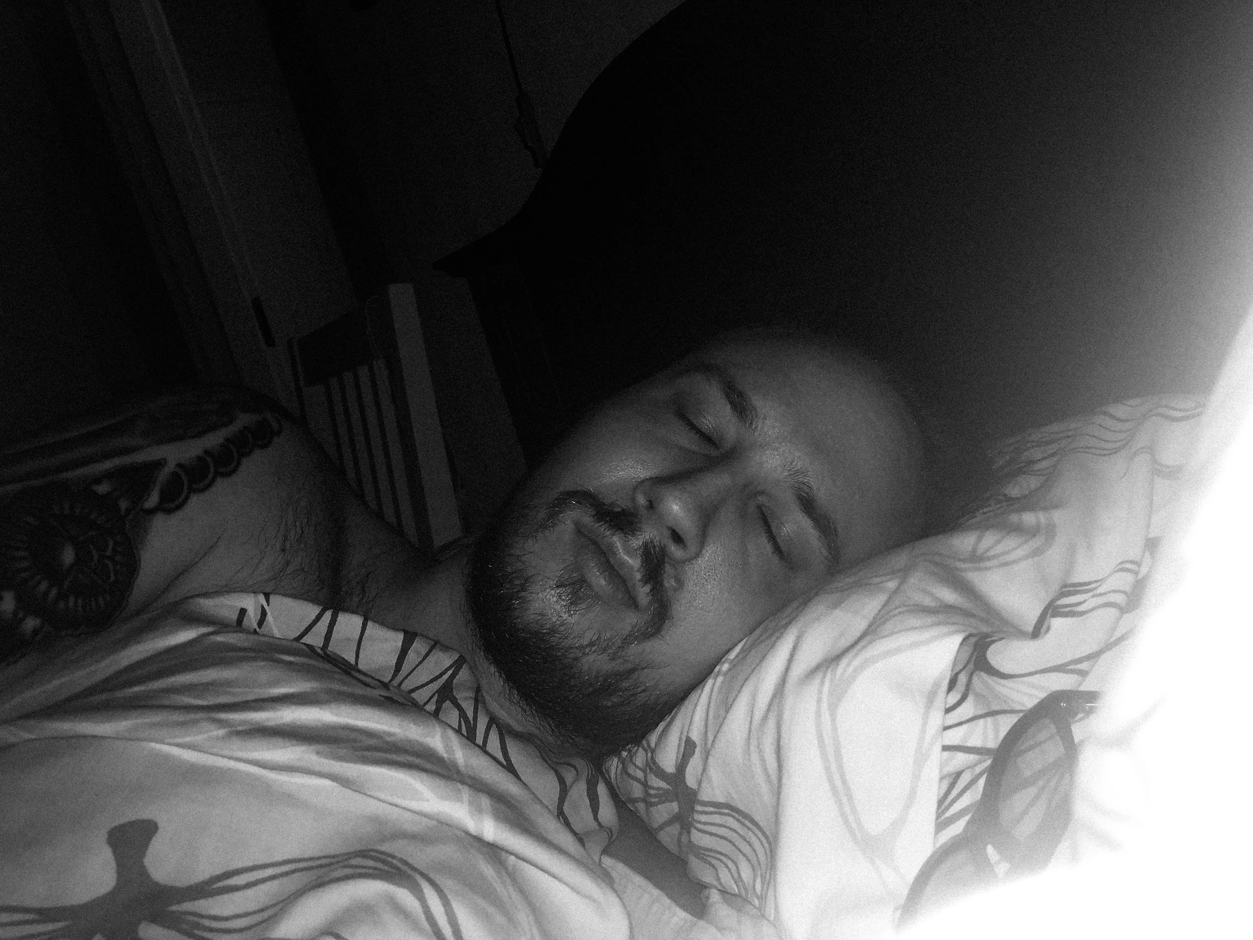 A black-and-white photo of Niko resting his head on a pillow, his eyes closed, and he’s under a blanket; the camera’s flash lights him in an otherwise dark bedroom.