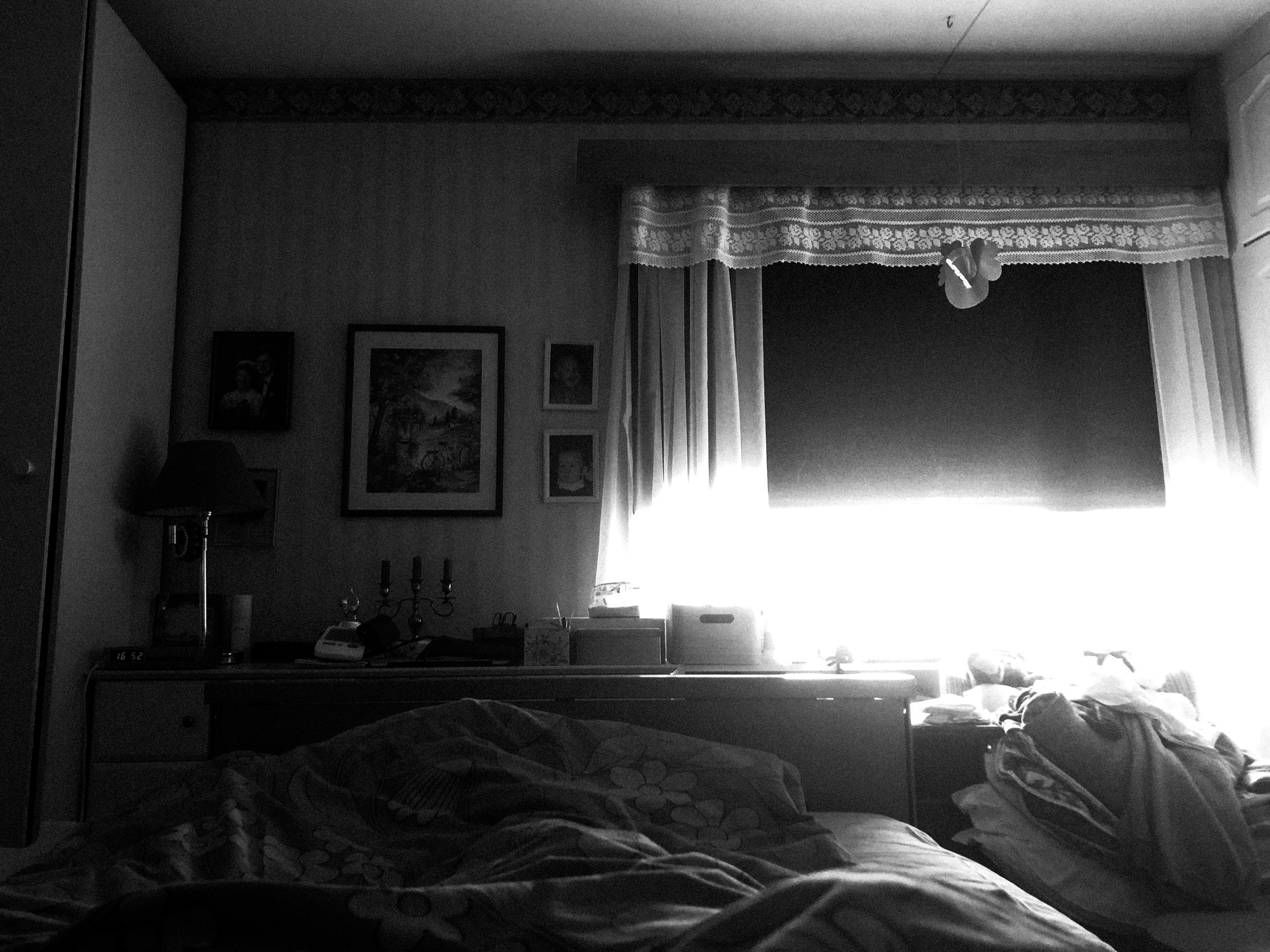 A black-and-white high-contrast photo of a dark bedroom window; the blackout curtain is lifted a bit to let some light in, which looks like it’s glowing into the room.