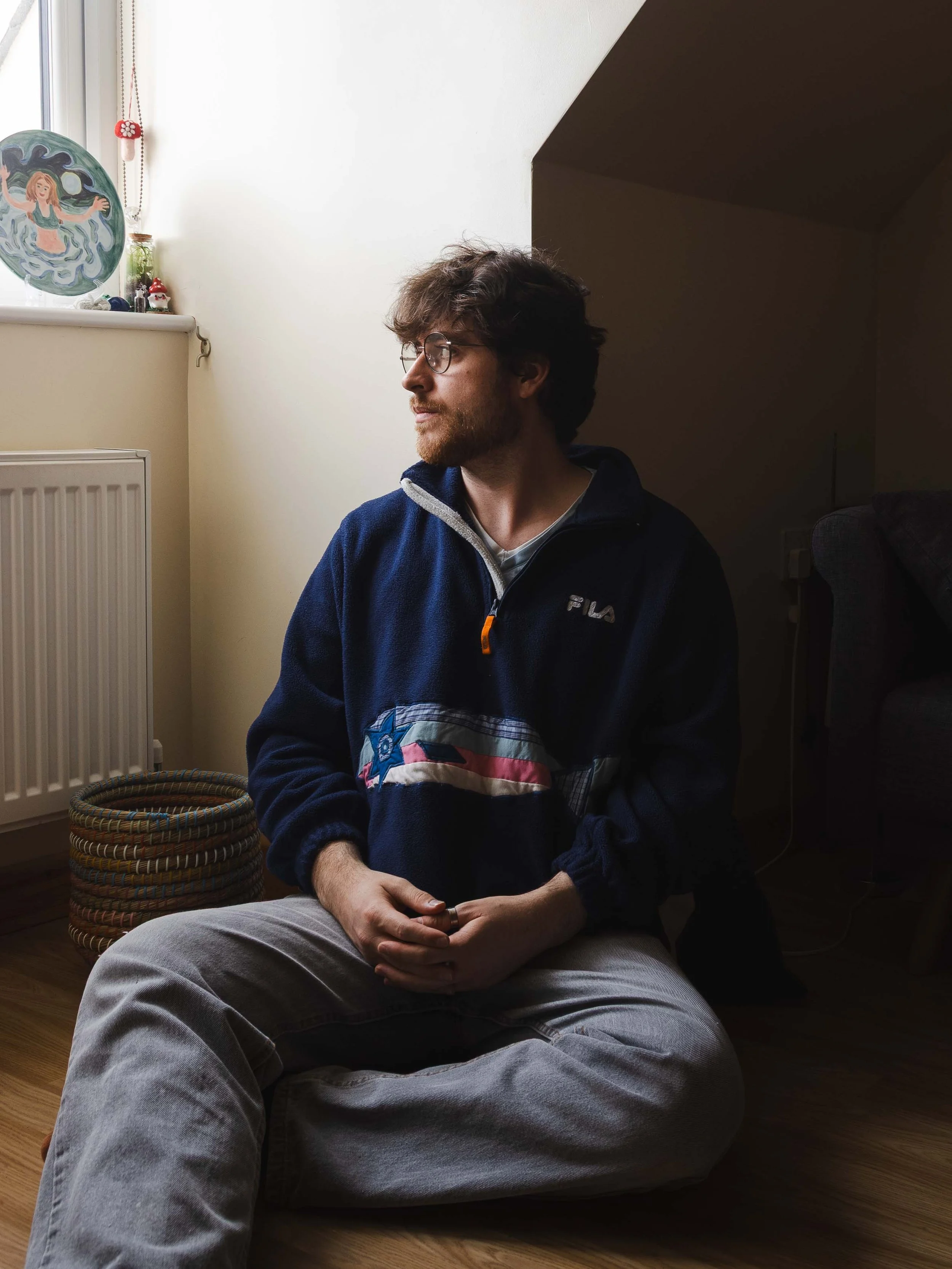 Will, a fair-skinned man with short curly brown hair, a beard and moustache, and round glasses, sits on the floor against a knee wall, looking to his right.