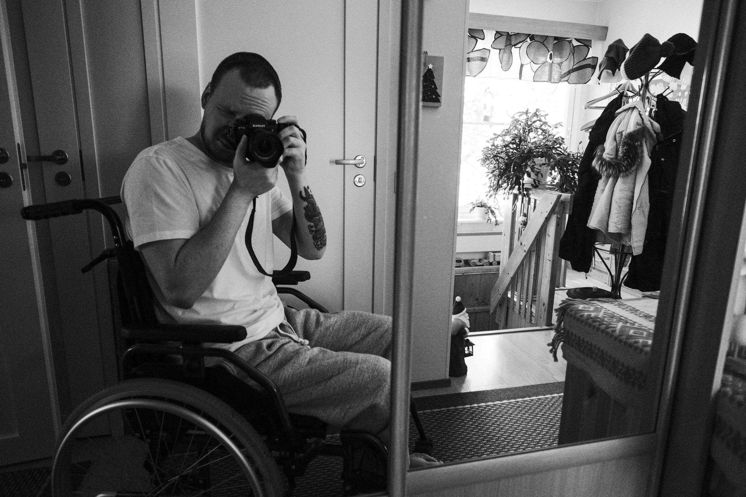 A black and white photo of Niko, with short hair and a beard, wearing a white t-shirt and grey sweatpants, sitting in a wheelchair. He takes a self-portrait through a cabinet’s mirrored door. A rack of clothes and a window are in the background.