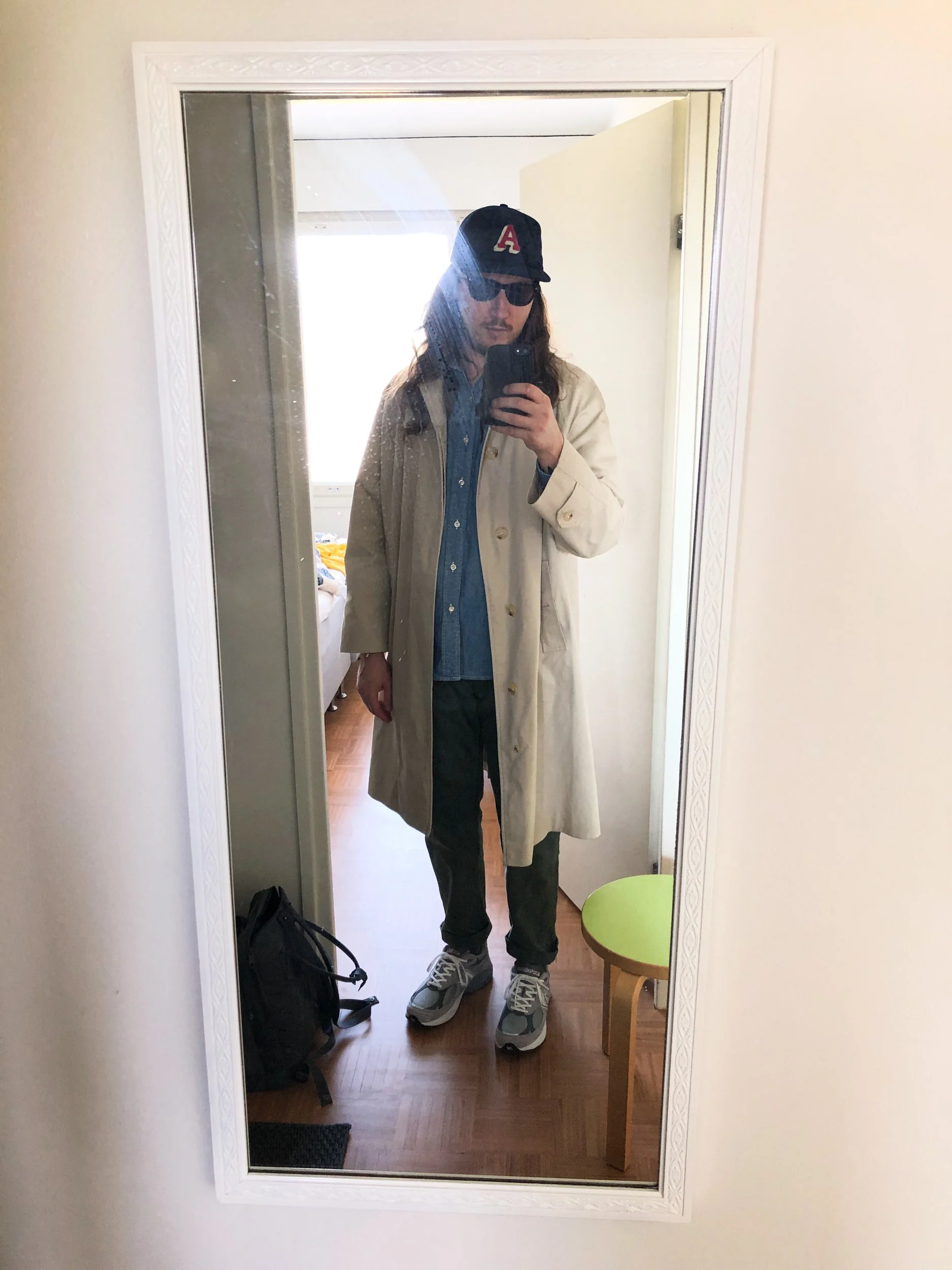 A colour photo of Niko, a man wearing a tan mac coat, a cap, sunglasses and sneakers, taking a selfie via a mirror.