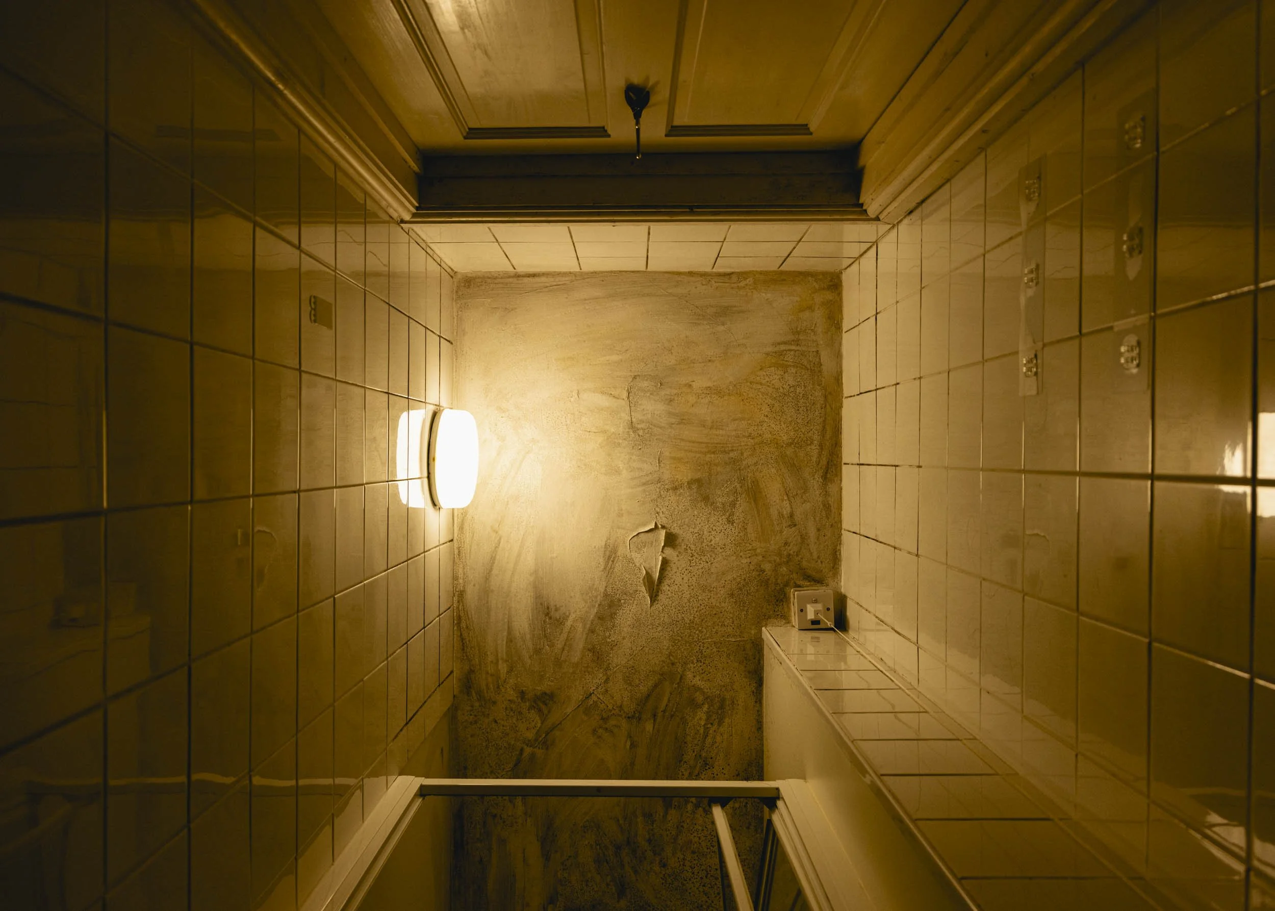A photograph of a ceiling in a narrow room streaked with black mould and cracked paint. The walls are covered in white square tiles, with a wooden door and a glass shower door. A round light on the left wall illuminates the space in a yellow glow.