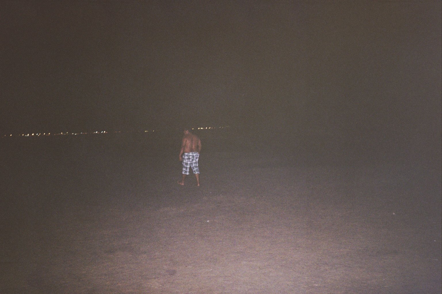 A hazy and dark grey coloured photograph of a person walking in the distance. In the far distance, there are some city lights.
