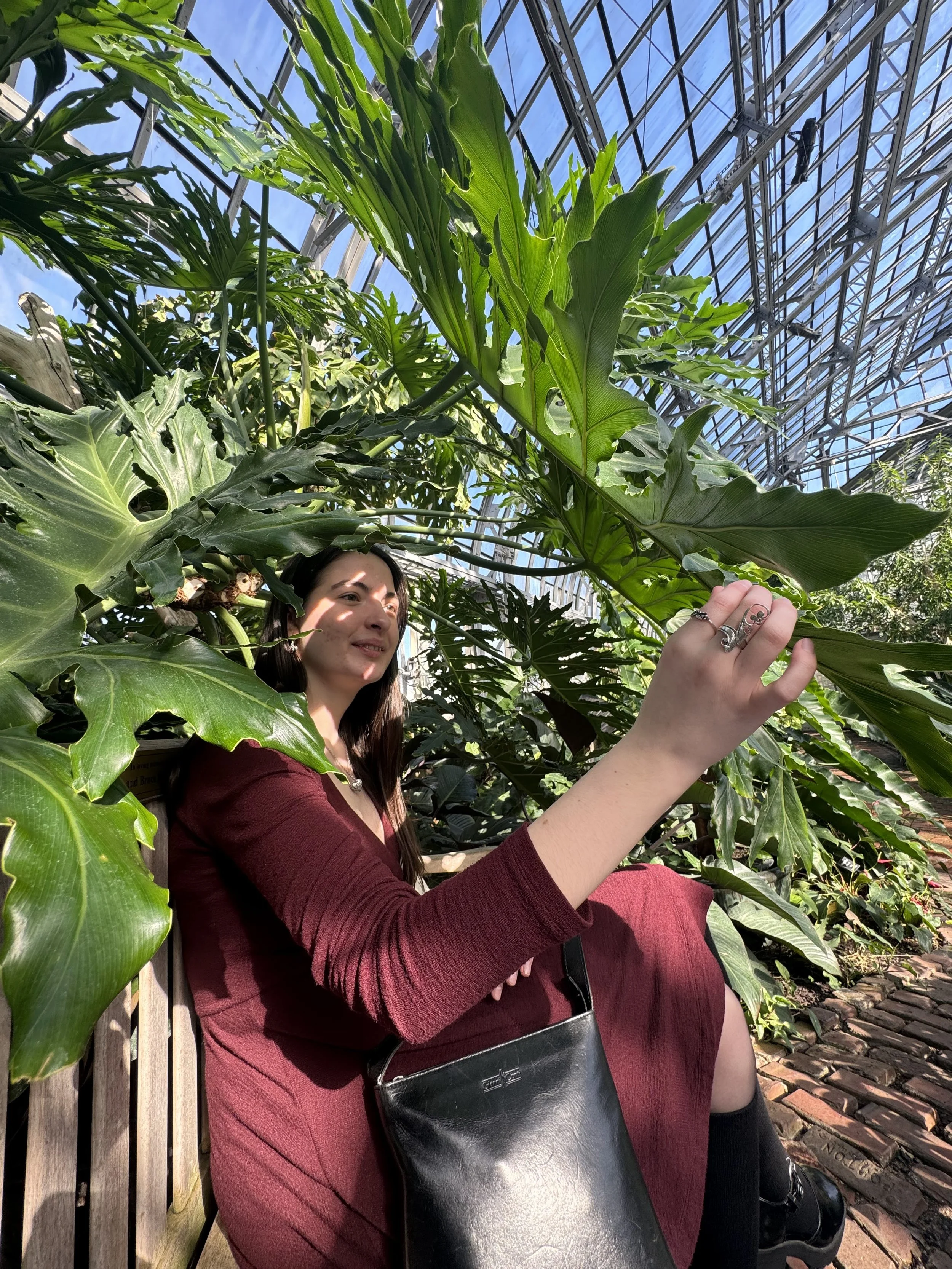 Lily, a young white woman with brown hair, is wearing a red dress. She is looking away from the camera and reaching their hand to touch a large leaf. She is surrounded by plants, the background filled with green leaves and trees.