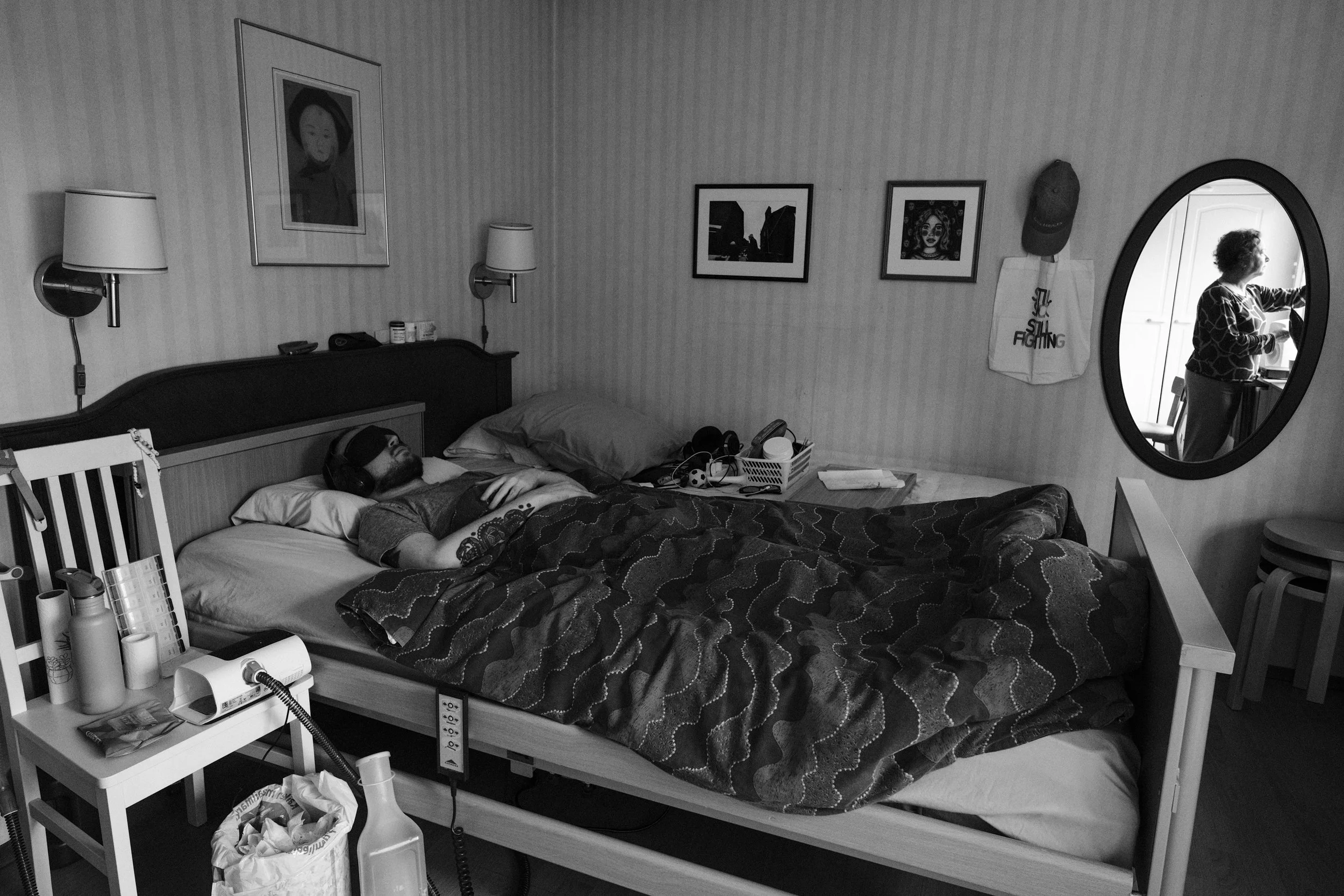 A black and white photo of a man lying on a hospital bed, hands crossed on his chest, under a duvet, and wearing an eyemask and earmuffs. The room has framed pictures and personal belongings. A mirror reflects a woman closing the curtains.