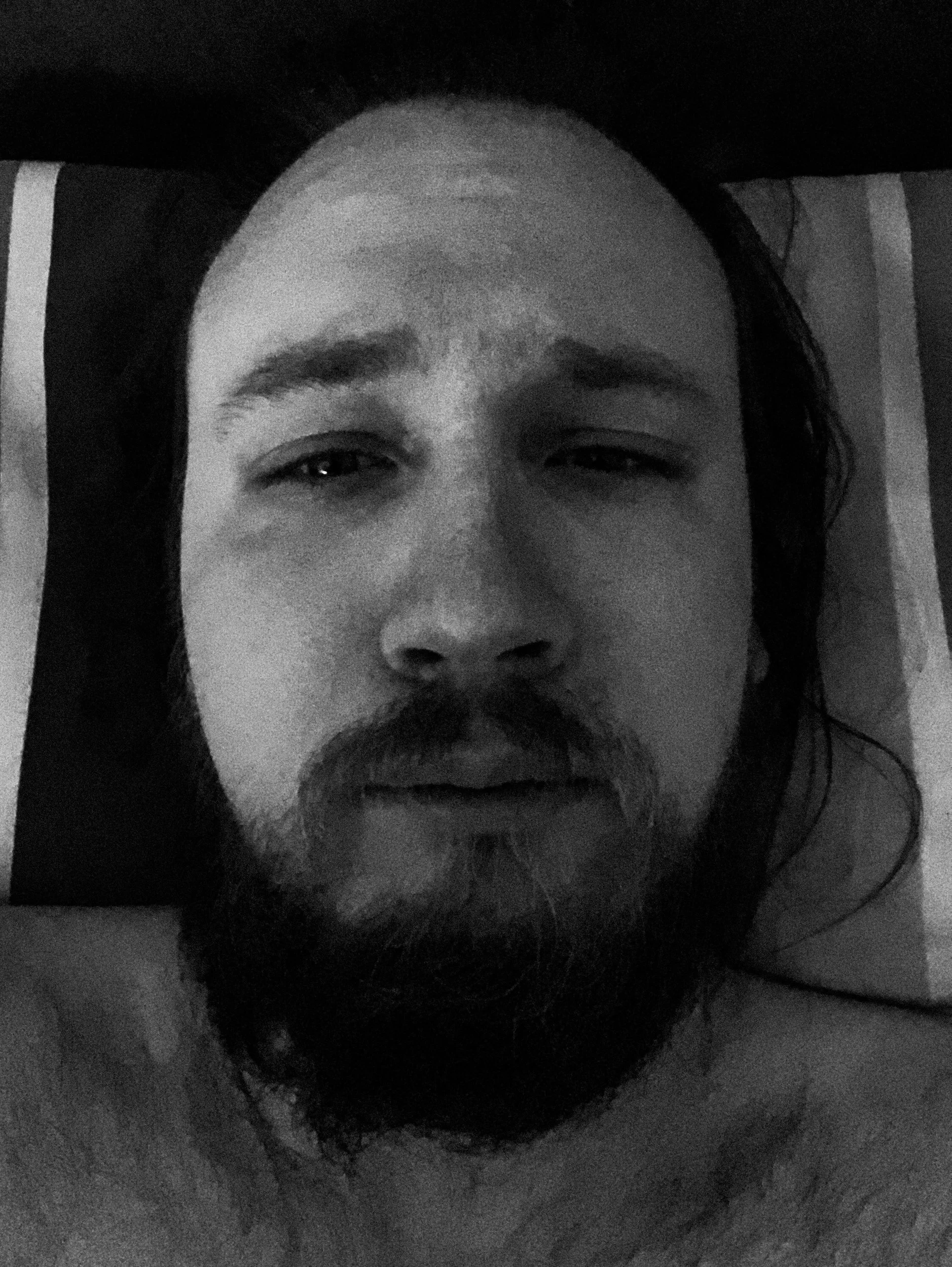 A black-and-white selfie photo of Niko in a dark bedroom; he’s looking directly at the camera with teary eyes while lying in bed; he has a messy beard and long hair.