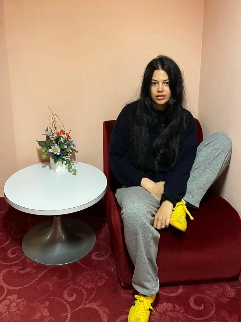 A young woman with very long black hair sits with one leg tucked up in a red velvet chair. She is wearing a long-sleeved back sweater, light grey sweatpants, and yellow sneakers. A round side table sits beside her with a small vase of flowers.