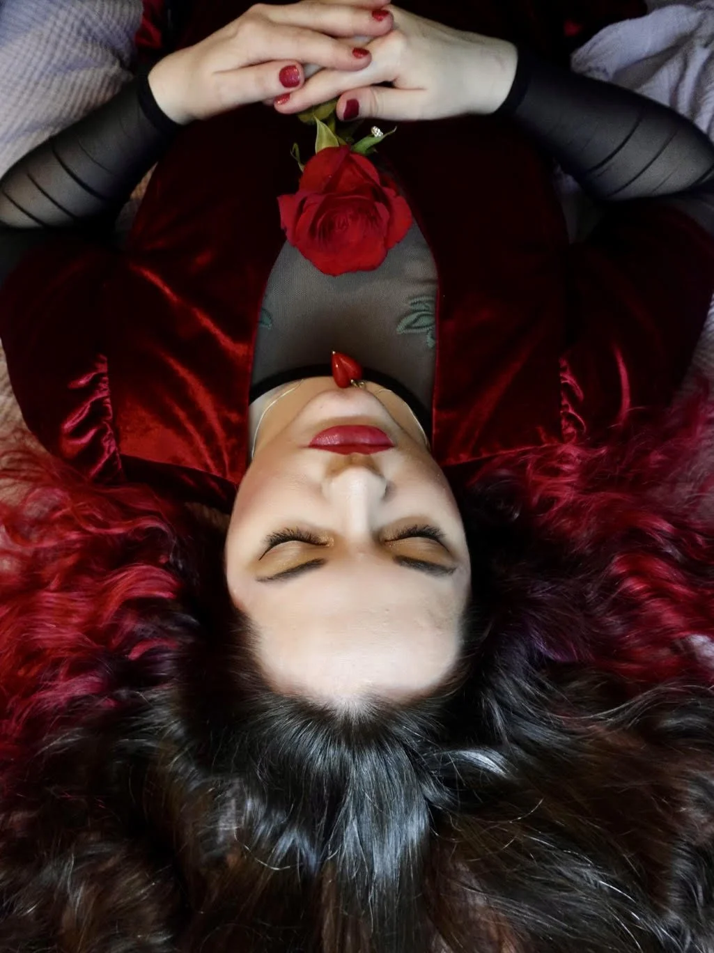 Fran, a young woman with fair skin and wavy brown hair dyed red in some parts, lying upside down while holding a red rose. She is wearing a red velvet dress with a black mesh undershirt.