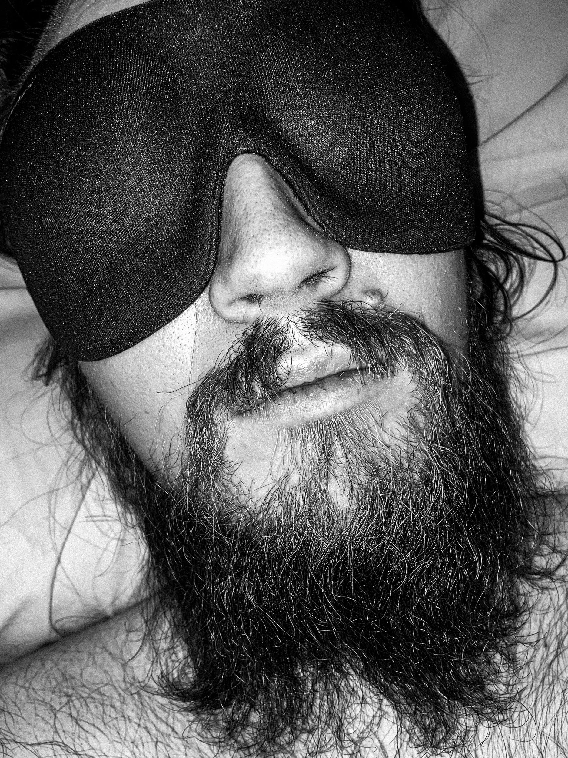 A black-and-white close-up photo of a shirtless Niko’s face, with a messy long beard, lying in bed with an eye mask on. The flash of the camera lights his face in the dark.