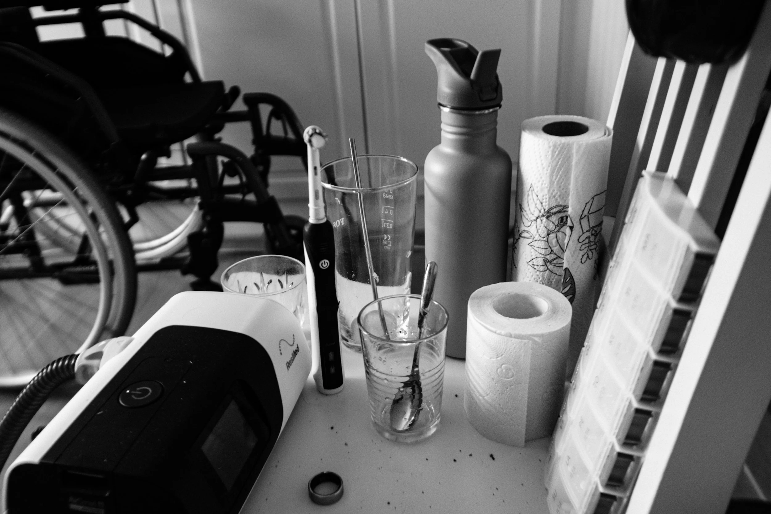 A black-and-white photo of a cluttered chair with an electric toothbrush, water bottle, paper towel roll, pill organiser, and other personal items. A wheelchair is in the background.