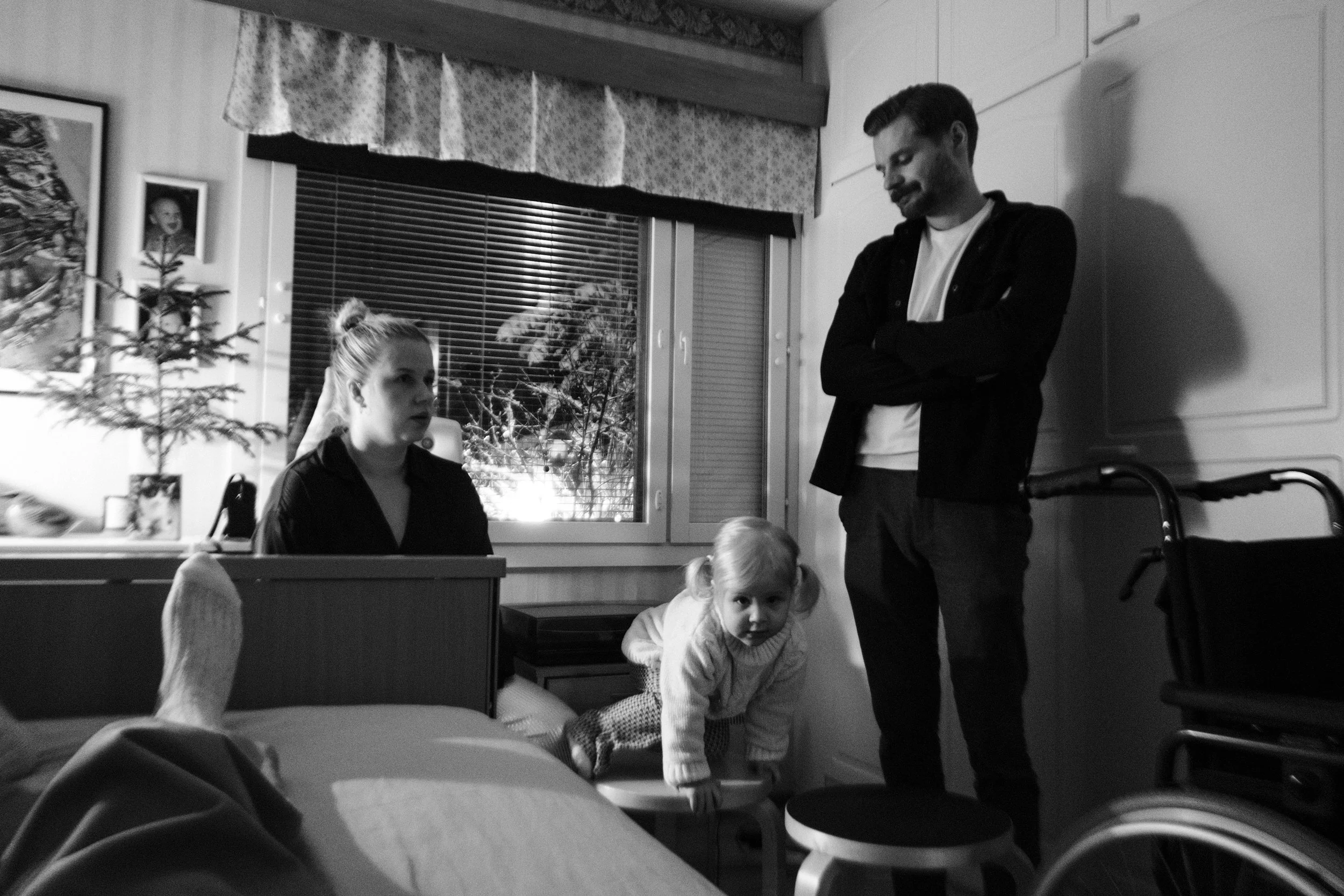 A black and white photo from the perspective of Niko lying in bed. A woman with a bun sits near the bed. A bearded man stands with arms crossed. A young child with pigtails climbs a stool between them. A wheelchair is in the background.