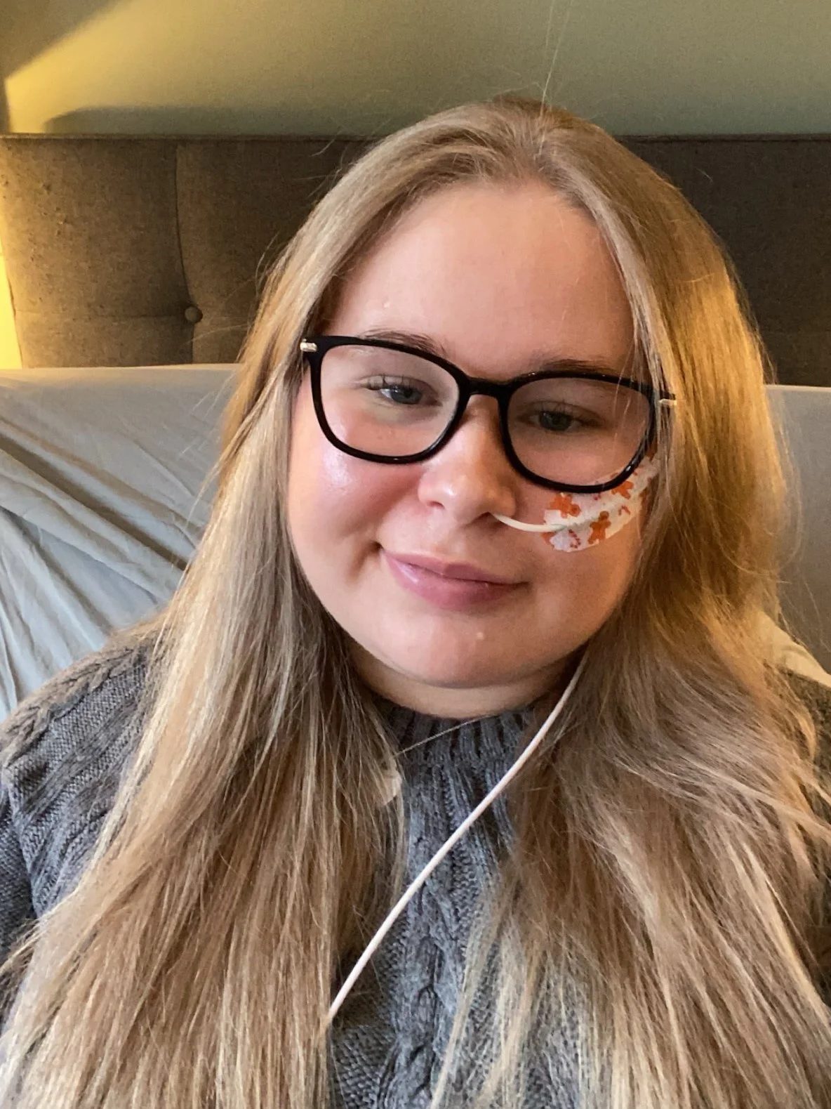 Katie, a young white woman with long blonde hair and glasses, wearing a gray cable-knit sweater. She has a nasal feeding tube and is seated in bed, looking at the camera with a slight smile.