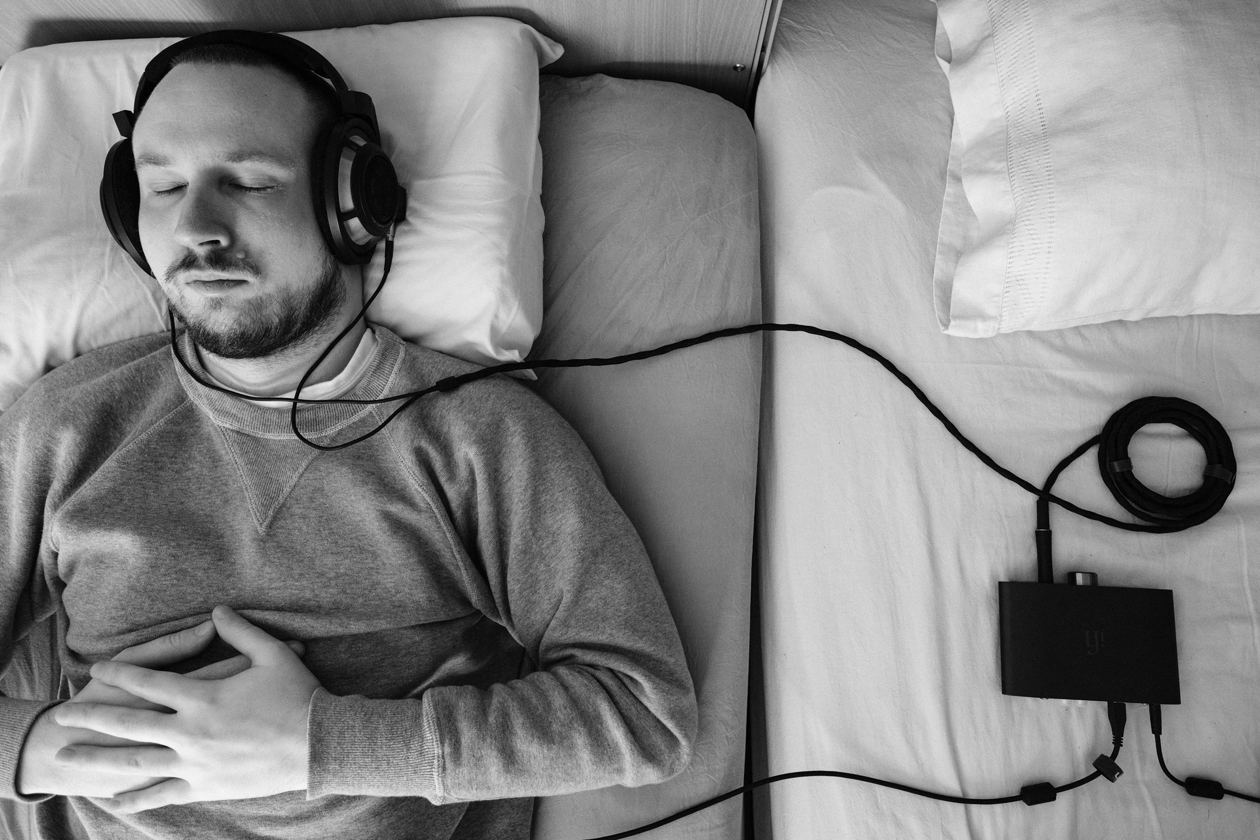 A black and white photo of Niko, with short hair and a beard, wearing a grey sweatshirt, resting in bed and listening to music eyes closed from wired headphones. Headphones are connected to a DAC/Amp. His hands are resting on his chest.