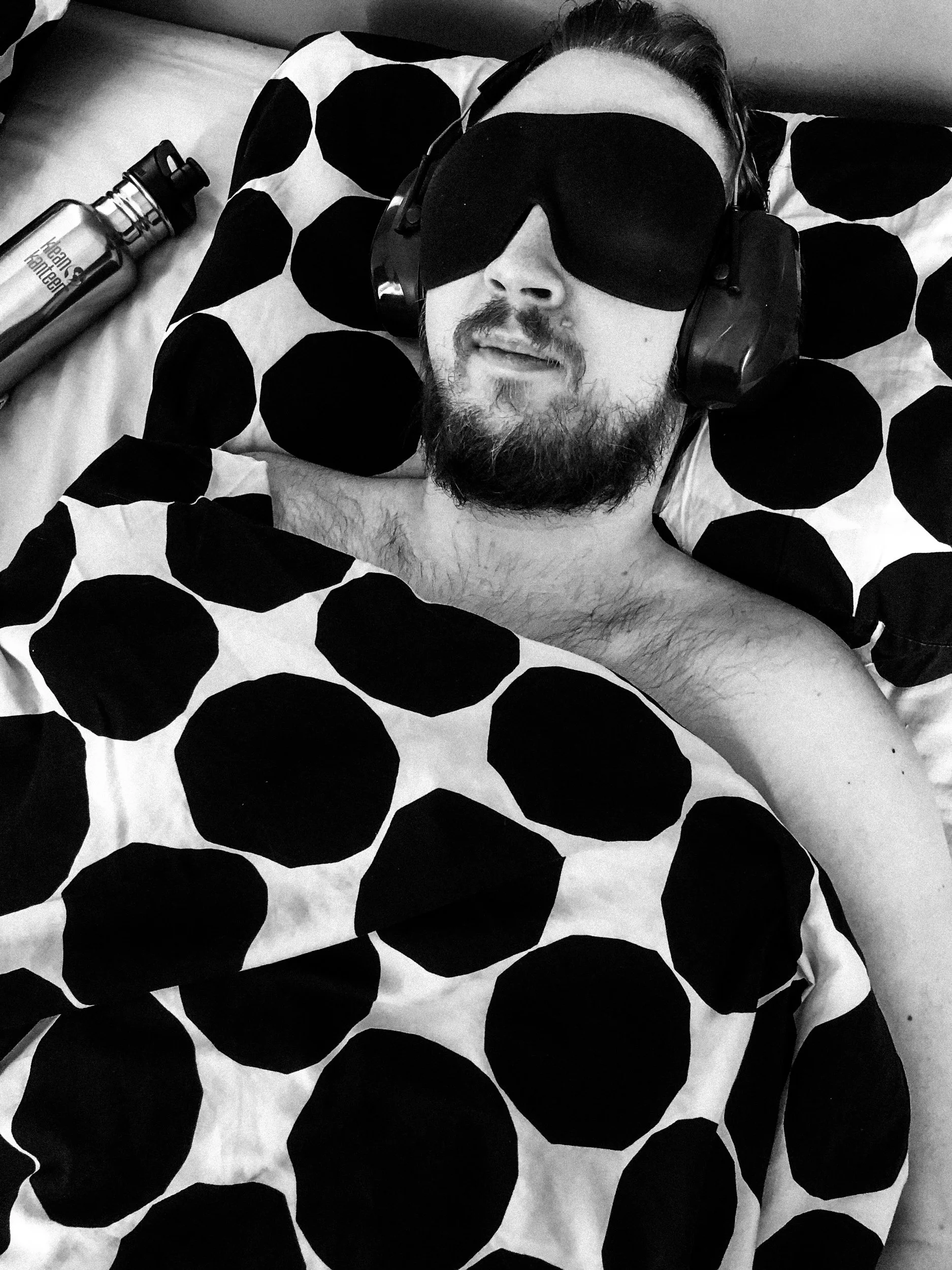 A black-and-white photo of Niko lying in bed with an eye mask and ear protectors on. He is tucked in under a blanket and has a water bottle next to him.
