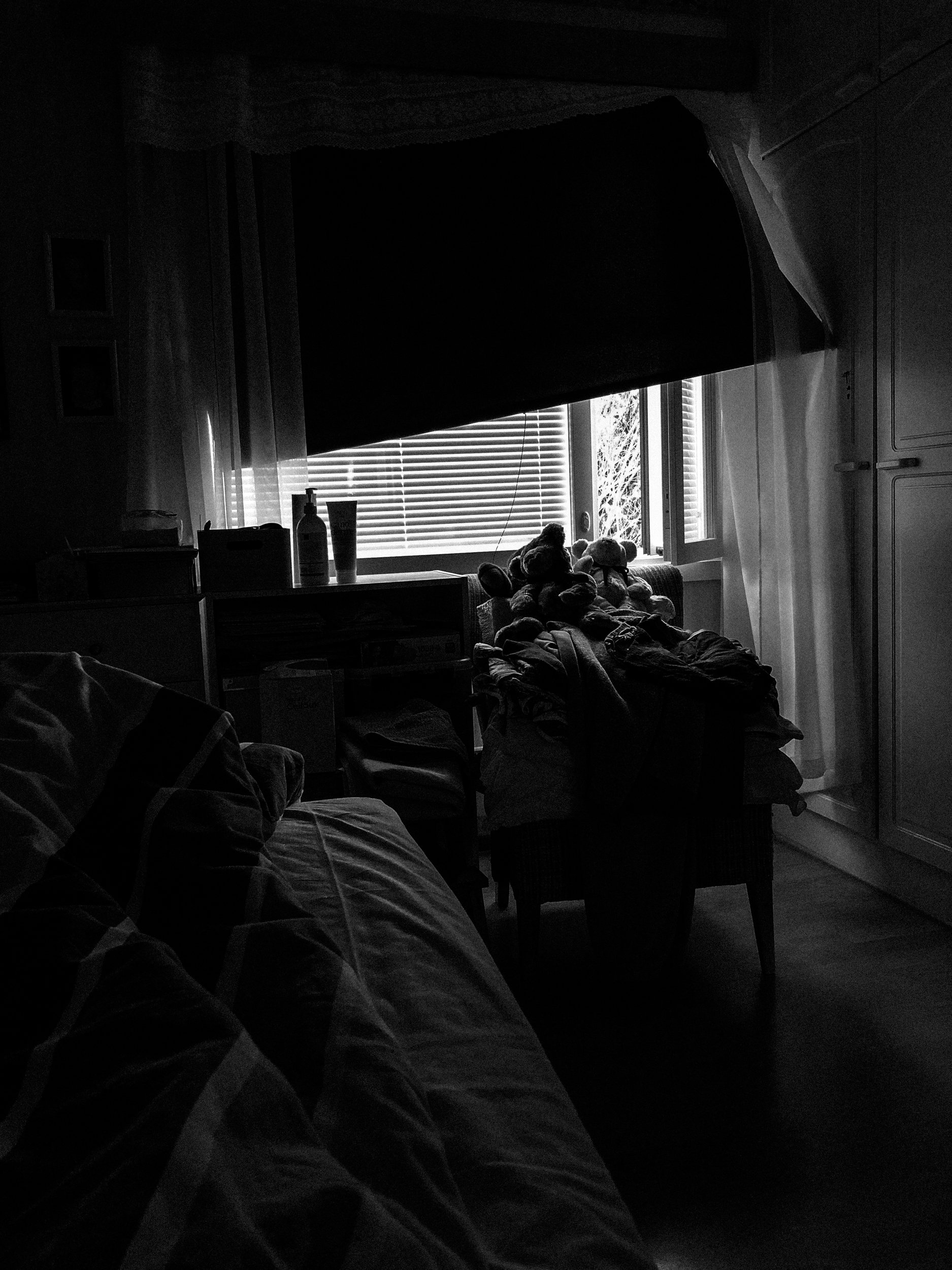 A black-and-white photo of Niko’s view from his dark bedroom. A small window is open, and the blackout curtain is lifted a bit, bringing some light to the room.