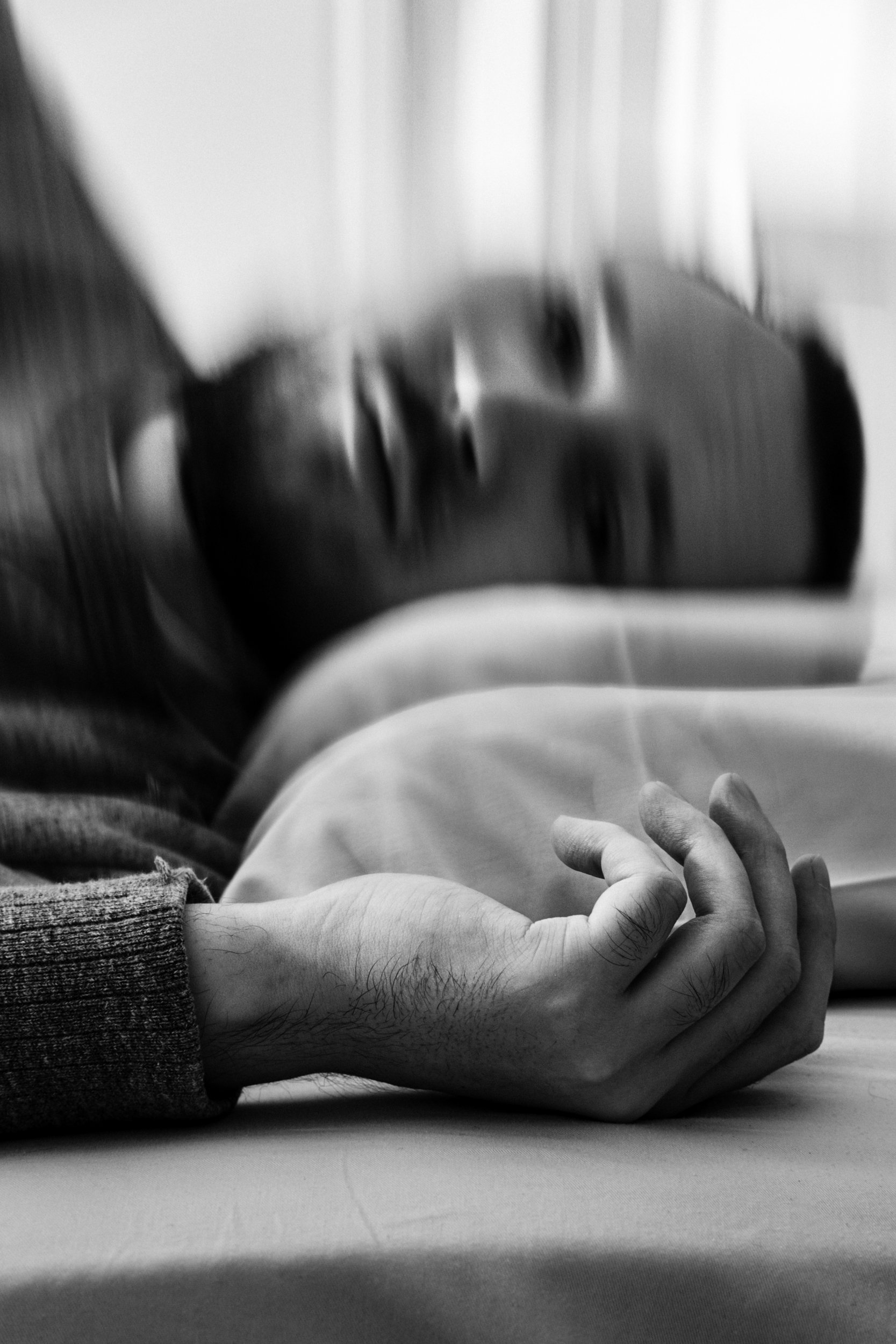 A black-and-white photograph of a man, Niko, resting his head on a pillow. His hand is in the foreground laying against the bed. His face appears blurry and distorted in the background.
