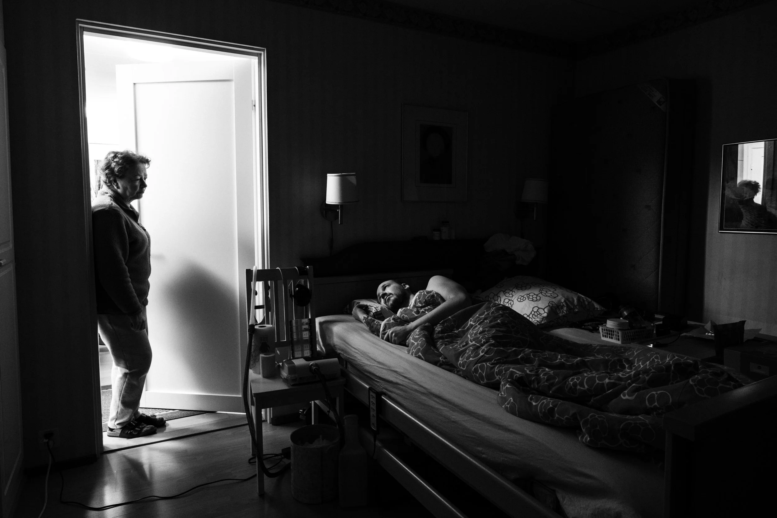 A black-and-white photo of a dimly lit bedroom. A man with a shaved head and beard, lies in bed under a blanket, looking at Päivi, who stands in the doorway, lit by another room. A chair beside the bed holds medical equipment and personal items.