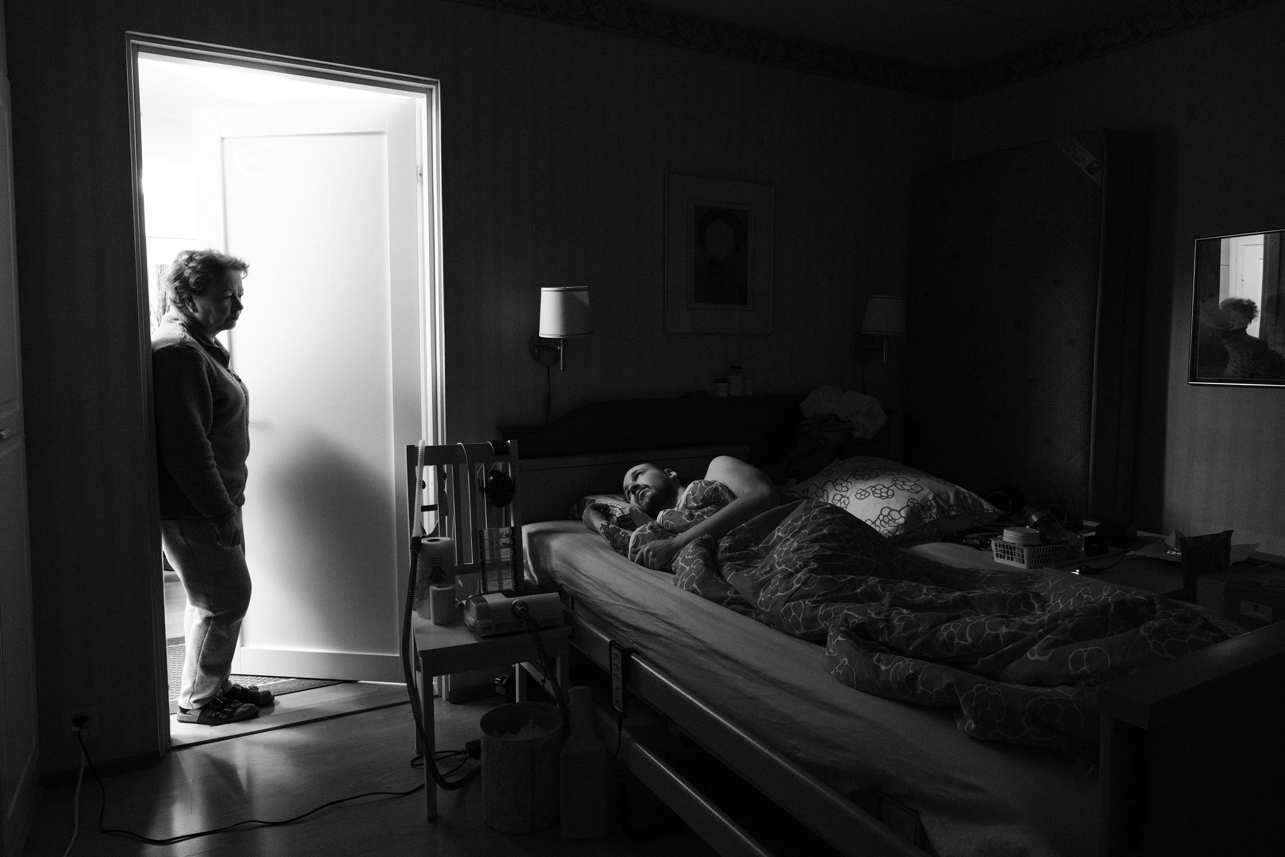 A black and white photo of a dimly lit bedroom. A man with a shaved head and beard, lies in bed under a blanket, looking at Päivi, who stands in the doorway, lit by another room. A chair beside the bed holds medical equipment and personal items.
