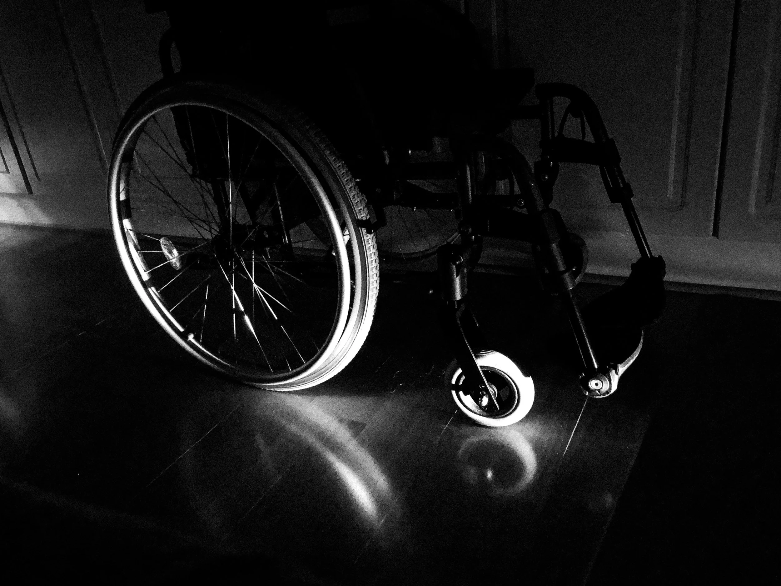 A black-and-white photo of a wheelchair in a dark room, only some parts of it are lit by light.