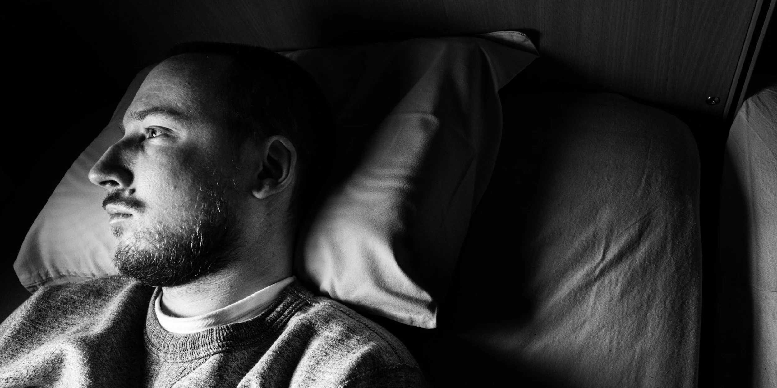 A black-and-white close-up of Niko resting his head on a pillow, gazing outside the frame. High-contrast lighting highlights his facial features, including his Roman nose, while the rest of the environment remains dark.