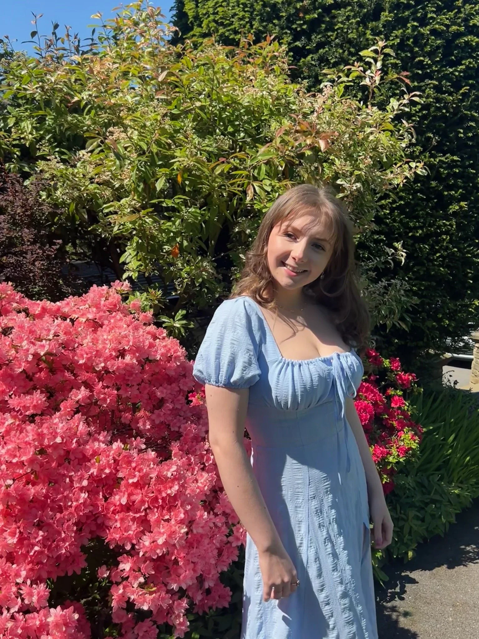 Emily, a young white woman with wavy brown hair wearing a pale blue short-sleeved dress, is standing in the sunlight in front of bushes and bright pink flowers and smiling.