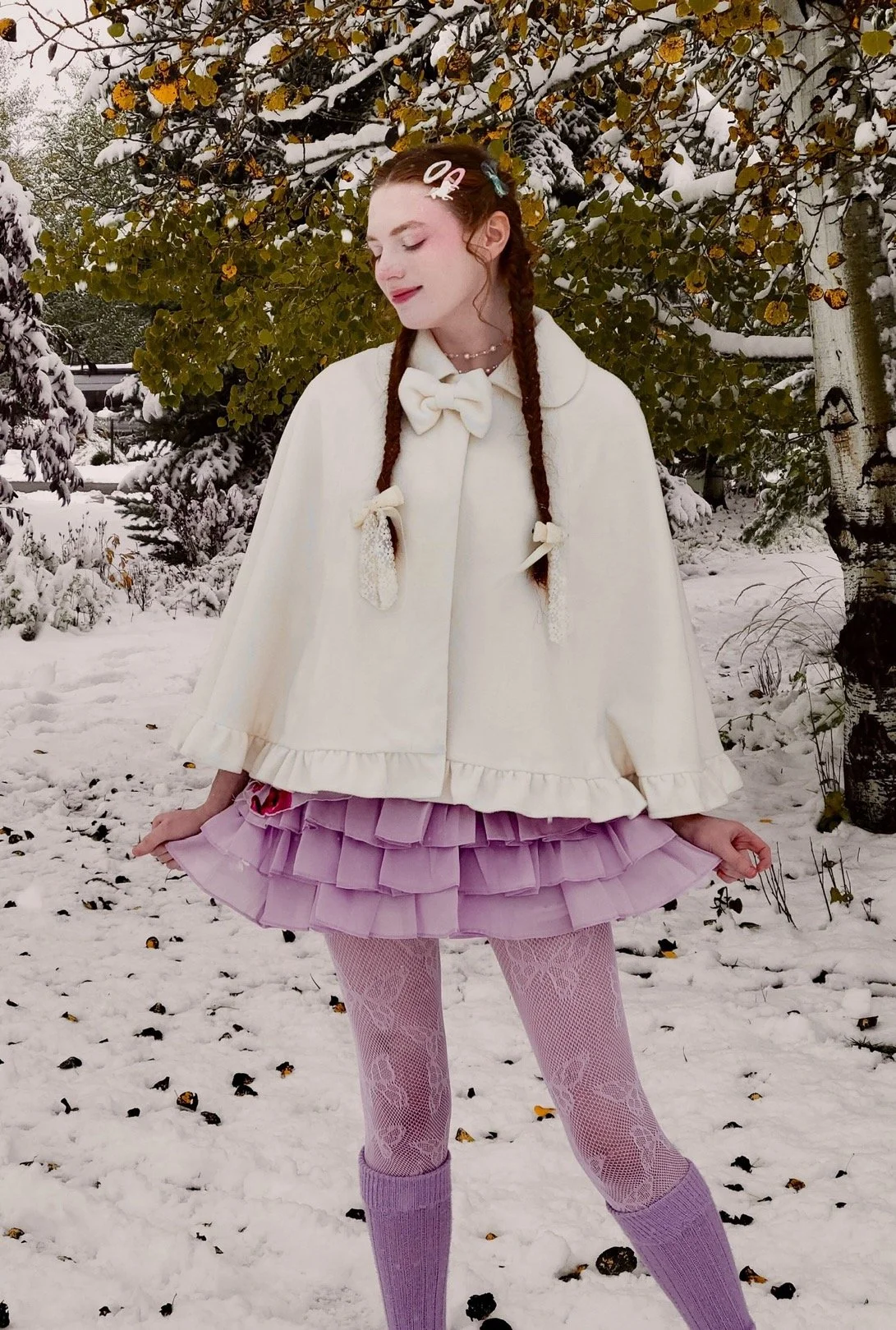 Lumie Mae, a feminine person with fair skin, stands in a snowy garden. Her red hair is in two braids, decorated with bows and barrettes. They wear a purple and white outfit with a cape, frilly skirt, butterfly tights, and tall socks.