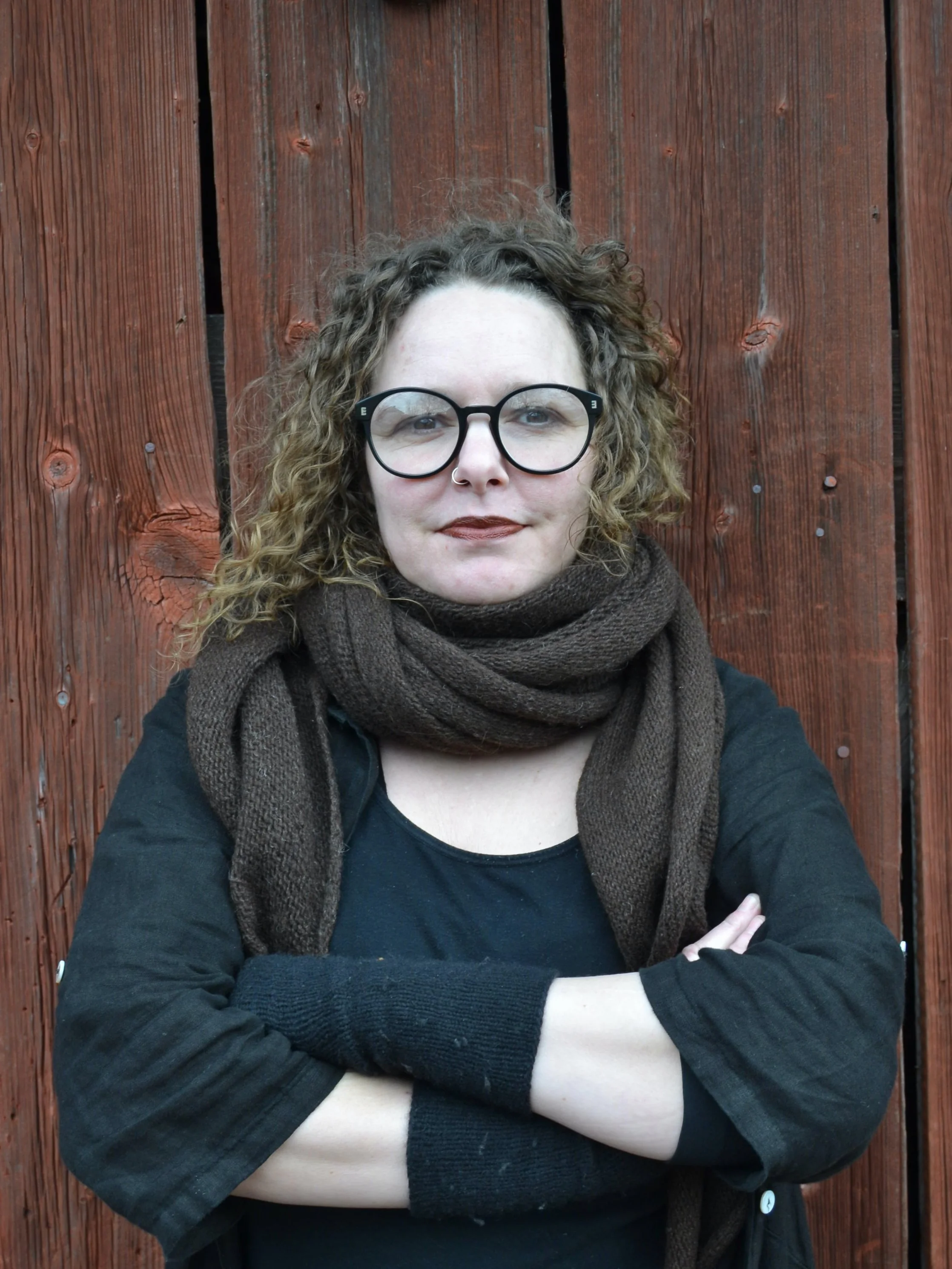Jenny, a white woman with curly brown hair and glasses, is standing in front of a wooden wall, looking straight at the camera with her arms crossed.