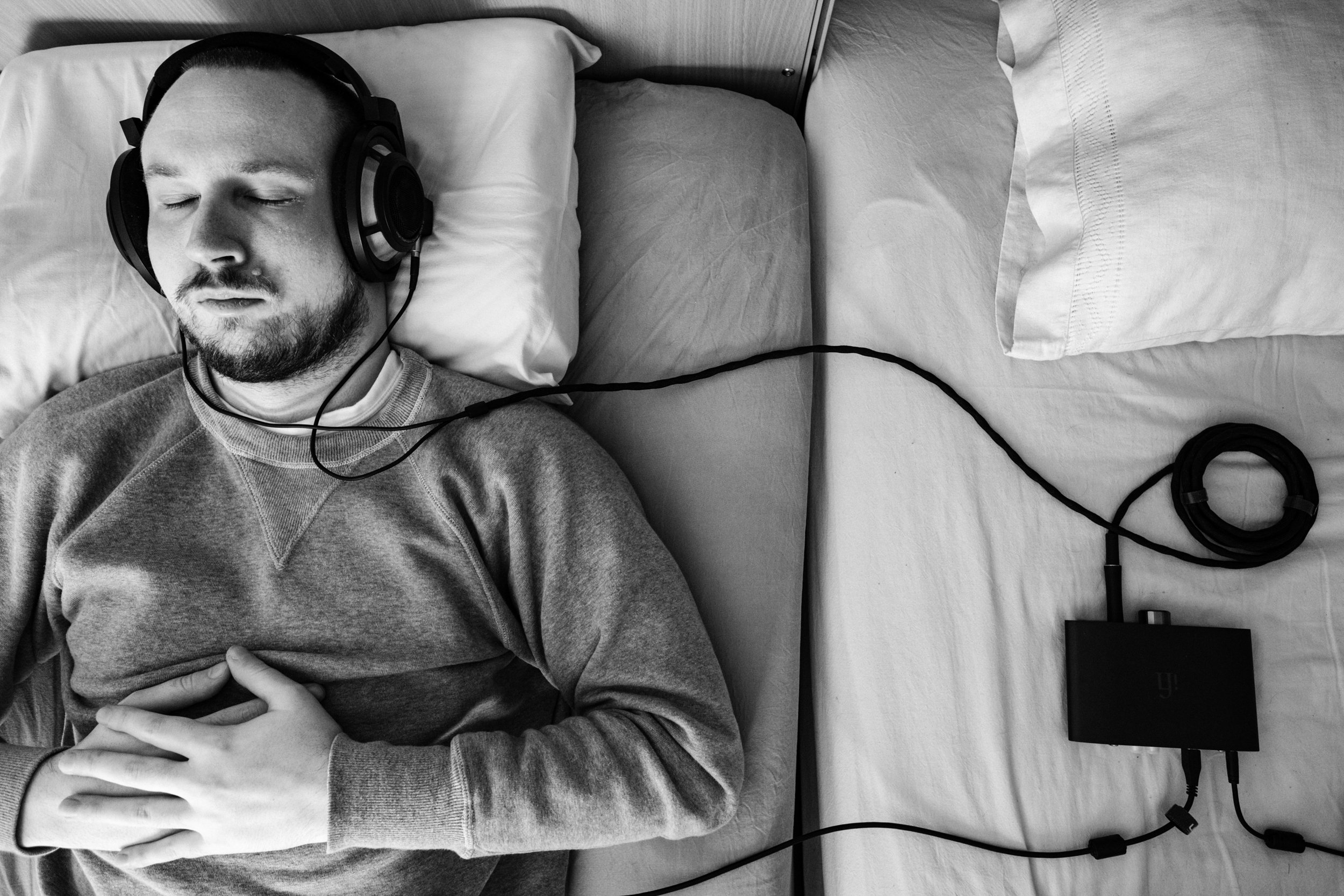 A black-and-white photo of Niko, with short hair and a beard, wearing a grey sweatshirt, resting in bed and listening to music eyes closed from wired headphones. Headphones are connected to a DAC/Amp. His hands are resting on his chest.