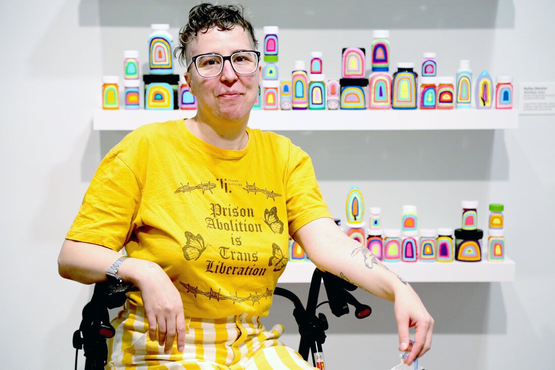 A half-length portrait of a person seated on a walker. Ze has short brown hair, glasses, arm tattoos, and is wearing a yellow shirt that reads 'Prison Abolition is Trans Liberation,' and yellow striped pants.