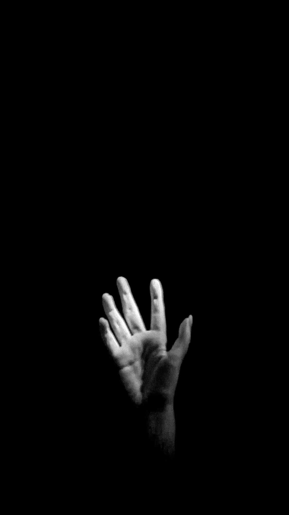 A black-and-white photo with a black background. There is a hand in the middle reaching upwards towards the light.