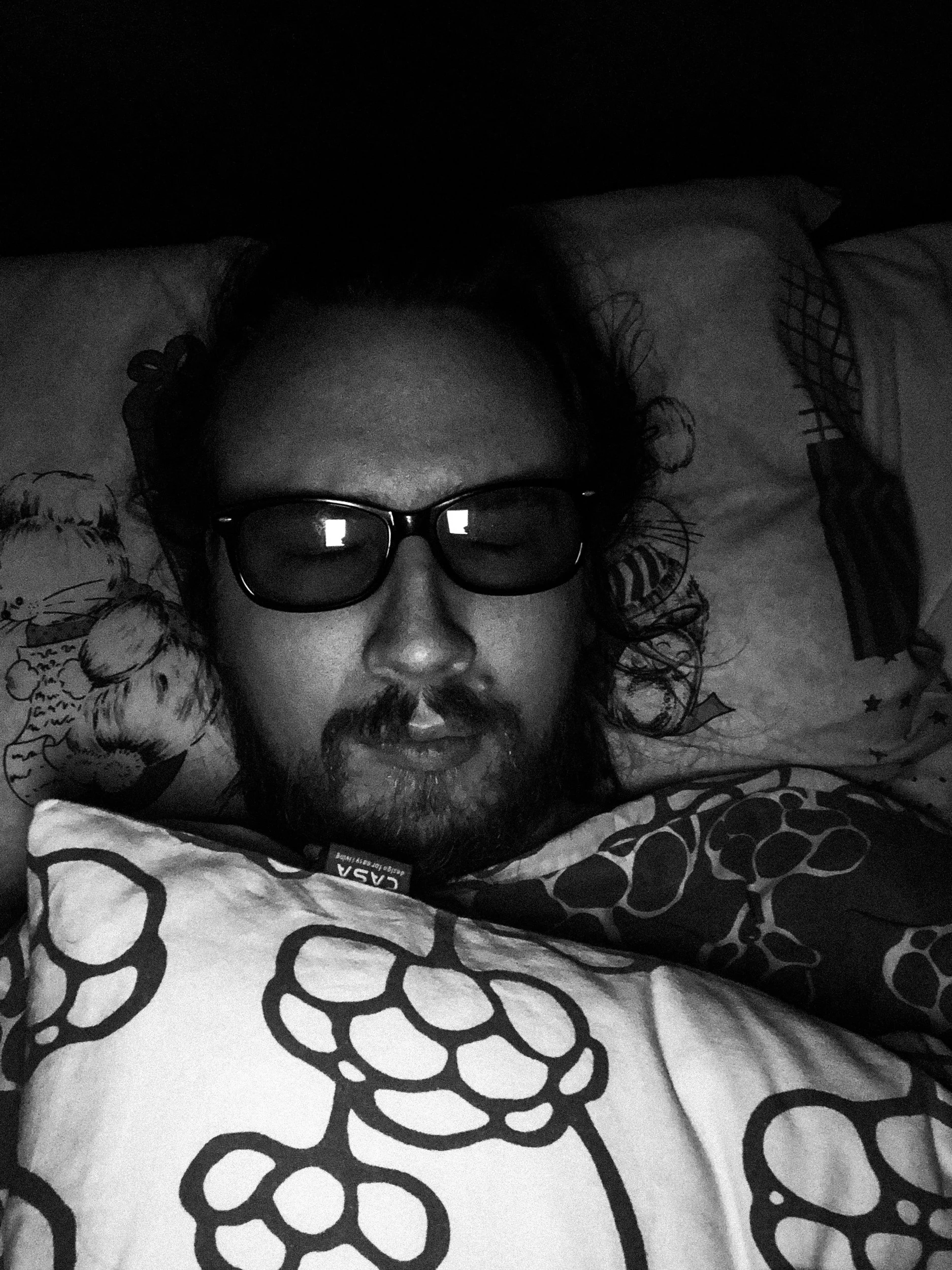 A black-and-white selfie photo of Niko in a dark bedroom; he has sunglasses on, and his eyes are closed; he has a messy beard and long hair. A pillow is resting on his chest.