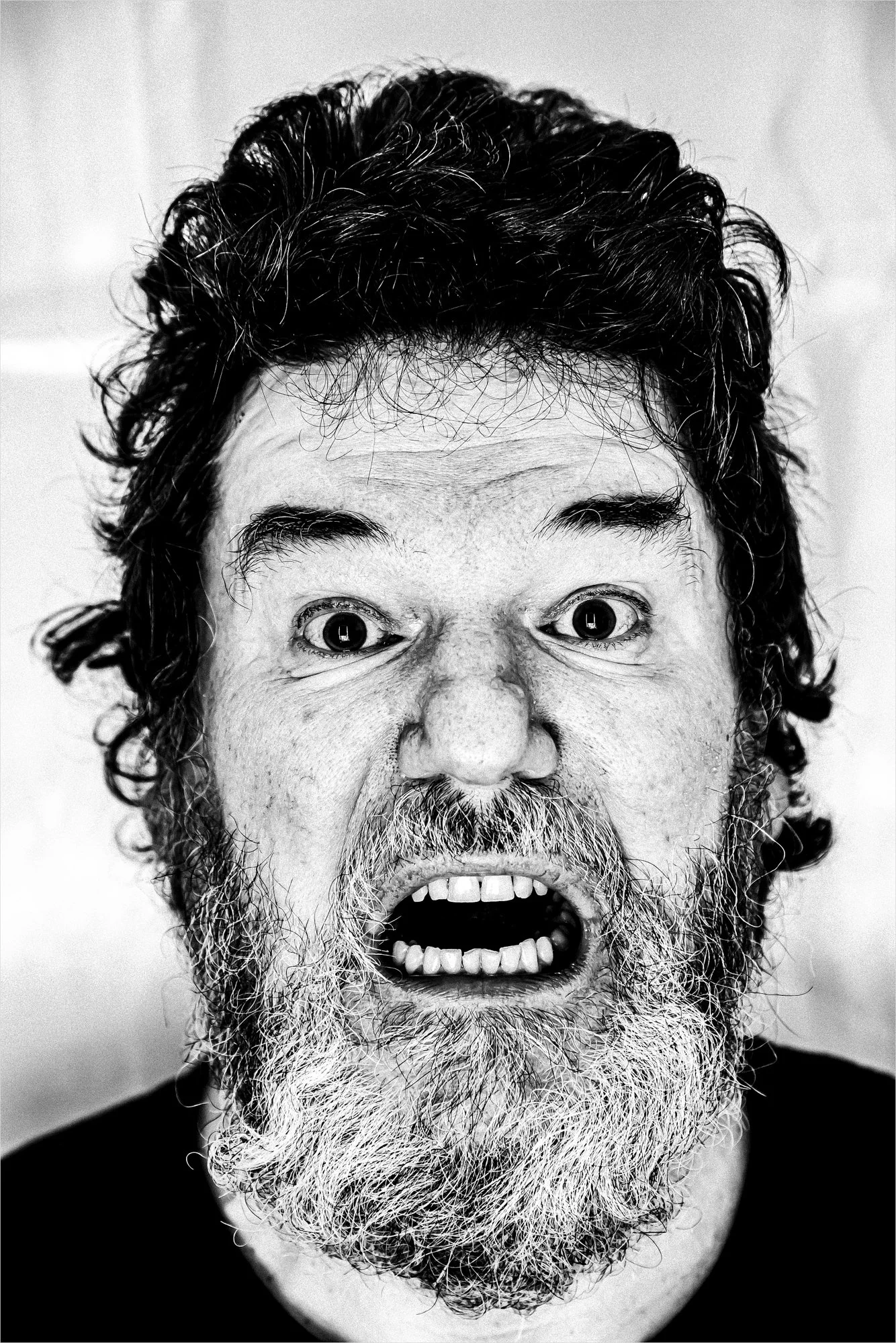 A black-and-white close-up photo of a screaming man with a longer beard and dark curly hair. He looks directly at the camera.