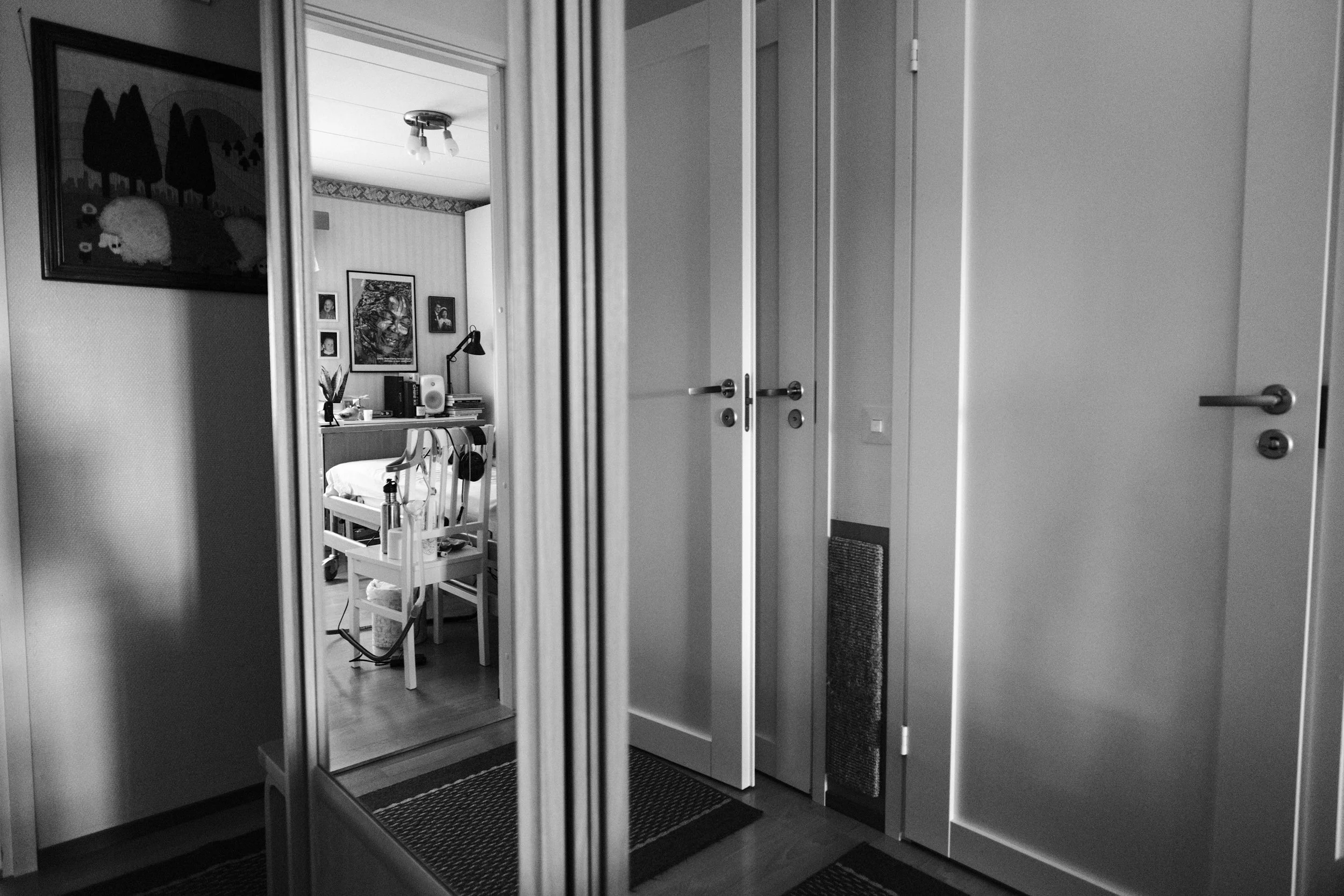 A black and white photo of a hallway’s cabinet with mirror doors. The mirrors reflect a bedroom seen from a doorway and multiple doors, which makes the image appear distorted and abstract.