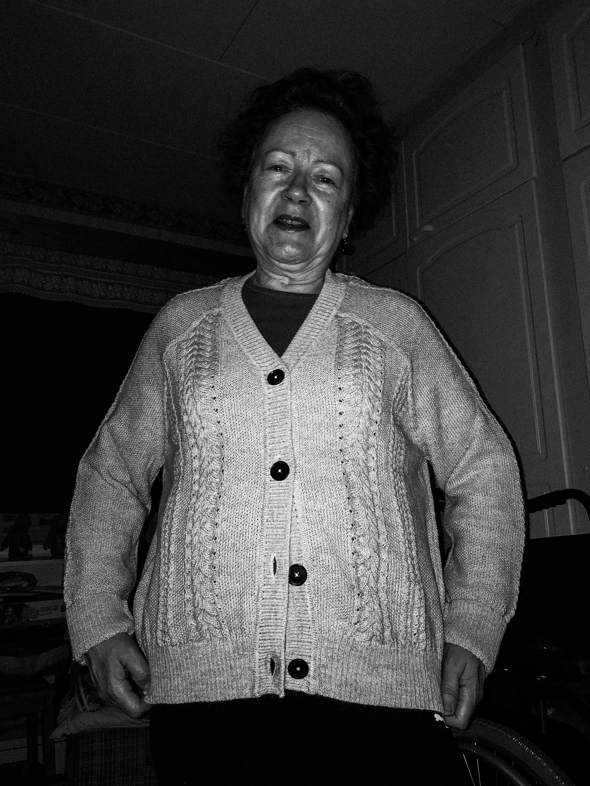A black-and-white photo of Niko’s mother, wearing a cardigan that she received as a gift. She is lit up by the camera’s flash and is laughing while looking at the camera.